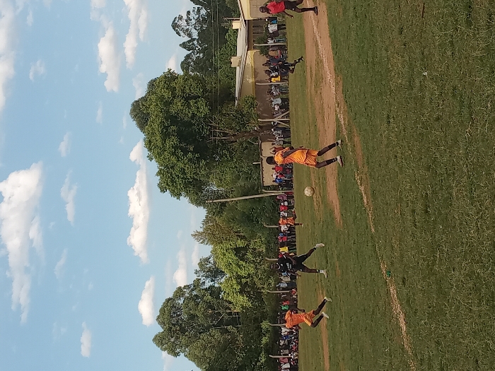 An outdoor football match is underway on a mixed grass and dirt field in Chevaywa ward, Kenya, beneath a light blue sky with scattered white cumulus clouds. Five male football players are visible. One player in an orange jersey and shorts runs from left to right in the foreground. Another player in a black jersey and shorts stands to his right, facing the center. A third player, also in an orange jersey and shorts, is positioned right of center, actively engaging with a light-colored football which is airborne near his feet. Two additional players, one in orange and one in red, are in motion further right and in the mid-background. A large crowd of spectators lines the background perimeter of the field, standing behind a fence. Beyond the spectators, dense green trees and light-colored buildings with dark roofs are present. A dark utility pole extends vertically on the right side of the scene.