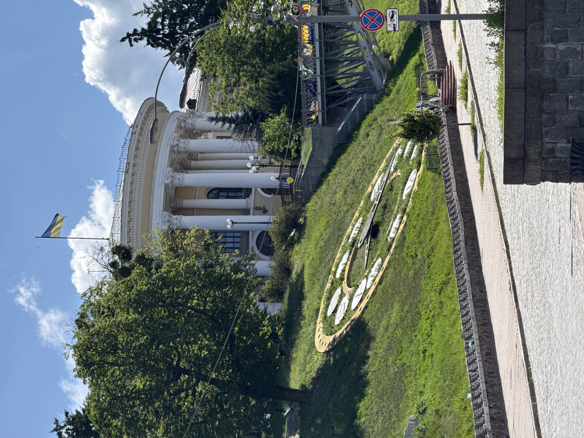 The image captures a view of the National University of Kyiv, also known as Shevchenko University, located in Kyiv, Ukraine. In the foreground, a large, decorative outdoor clock face is visible on a grassy slope. The clock is made of yellow material and white clock faces, with dark clock hands. A paved walkway with stone retaining walls borders the grassy area. Behind the clock, a metal bridge with intricate railings spans across a section of the slope. Above the bridge, stairs lead up towards the university building. The university itself is a grand, neoclassical structure with a curved facade, prominent white columns, and a Ukrainian flag flying from its roof. Lush green trees and foliage surround the building and the slope. The sky is blue with fluffy white clouds. In the lower right portion of the image, a traffic sign indicating no parking or stopping and a tow-away zone is visible next to a light post.