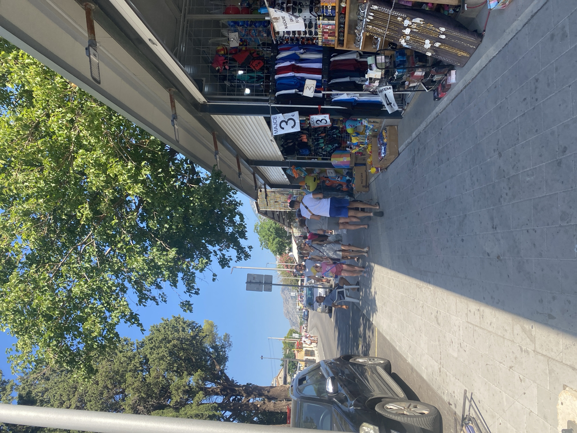 The image shows a street scene in Sutomore, Montenegro, on a sunny day. A black SUV is parked on the left side of the image, with a paved sidewalk in front of it. Several people are walking along the sidewalk, some are tourists. To the right of the sidewalk are several market stalls selling clothing, accessories, and souvenirs. The stalls have open fronts, displaying merchandise. In the background, there is a road with cars and more market stalls. Mountains can be seen in the distance. The atmosphere appears to be lively and commercial, typical of a tourist area.