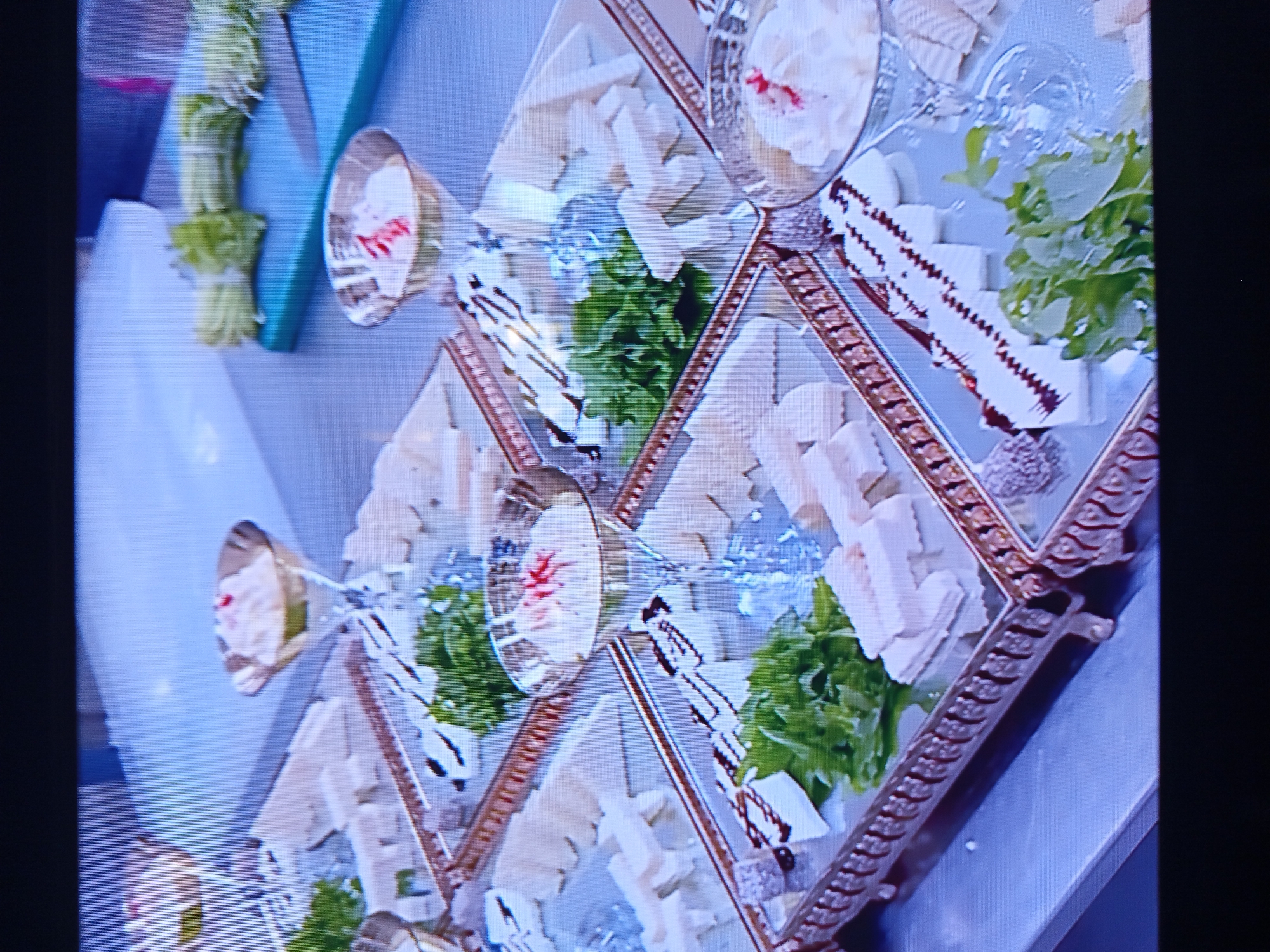 The image shows a close-up, angled view of several mirrored trays filled with cheese platters. The platters contain various types of cheese, some cut into rectangular blocks and others appearing as more solid pieces. There are also small bunches of green leafy herbs, likely for garnish, placed on the cheese. Some of the cheese is drizzled with a dark sauce, possibly balsamic glaze, creating a decorative pattern. In the background, out of focus, are several martini glasses filled with a white, creamy substance topped with a red garnish. To the right of the platters, a bunch of celery stalks tied with white twine is visible on a blue cutting board, along with a knife. The overall setting appears to be a kitchen or preparation area, with a stainless steel countertop. The image is a still from a video, indicated by the pixelation and slight blurriness. There are no people visible in the frame. The location in Pavlohrad, Ukraine is not discernible from the visual information provided in the image.