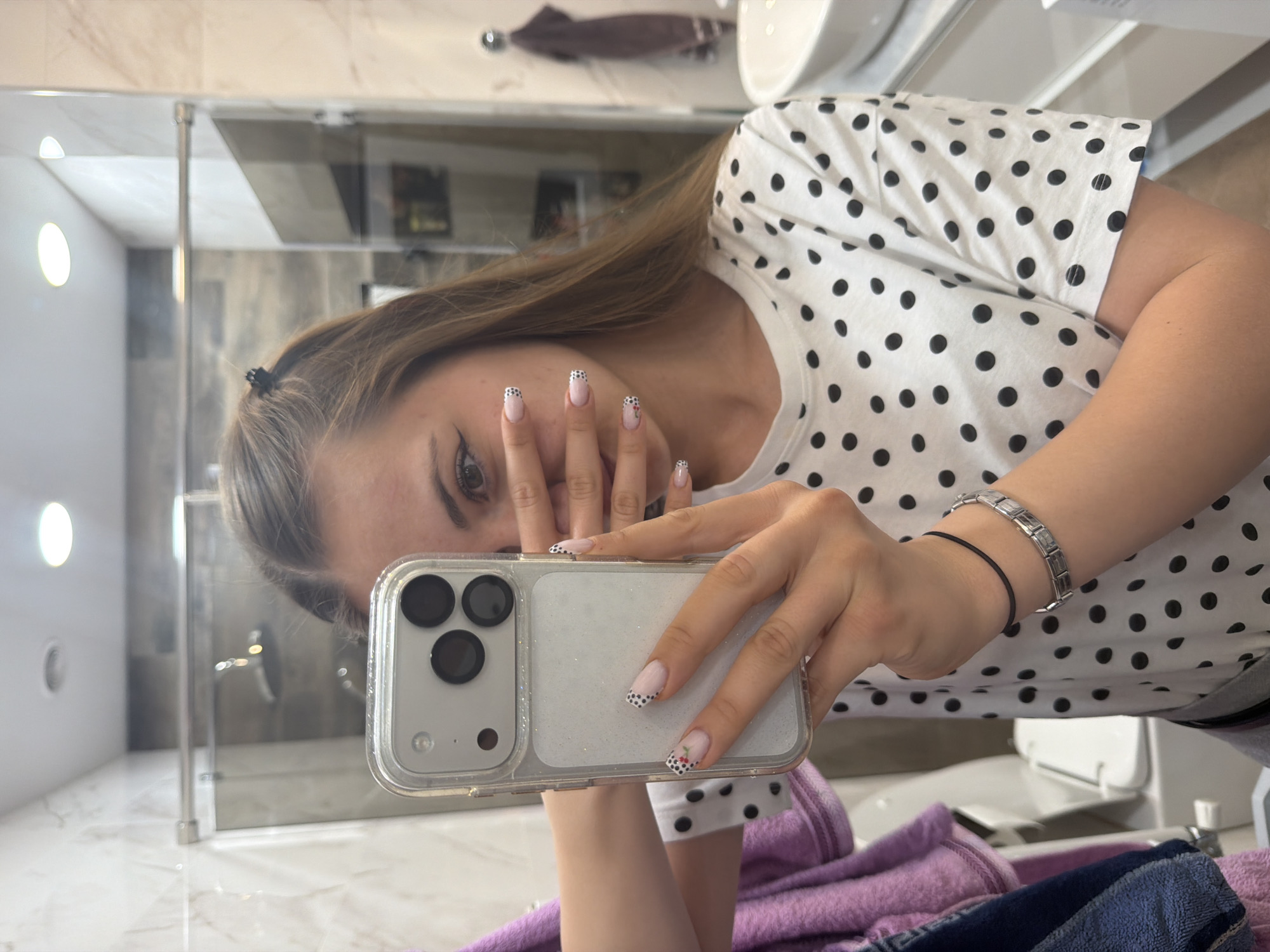 A woman is taking a selfie in a bathroom. The bathroom has white marble countertops, a glass shower enclosure, and a towel rack. The woman is wearing a white t-shirt with black polka dots. She is holding her phone up to her face, and her nails are visible. The nails are painted with a French tip and black polka dots. She is also wearing bracelets. The location is within a building in Buhovo, Bulgaria.