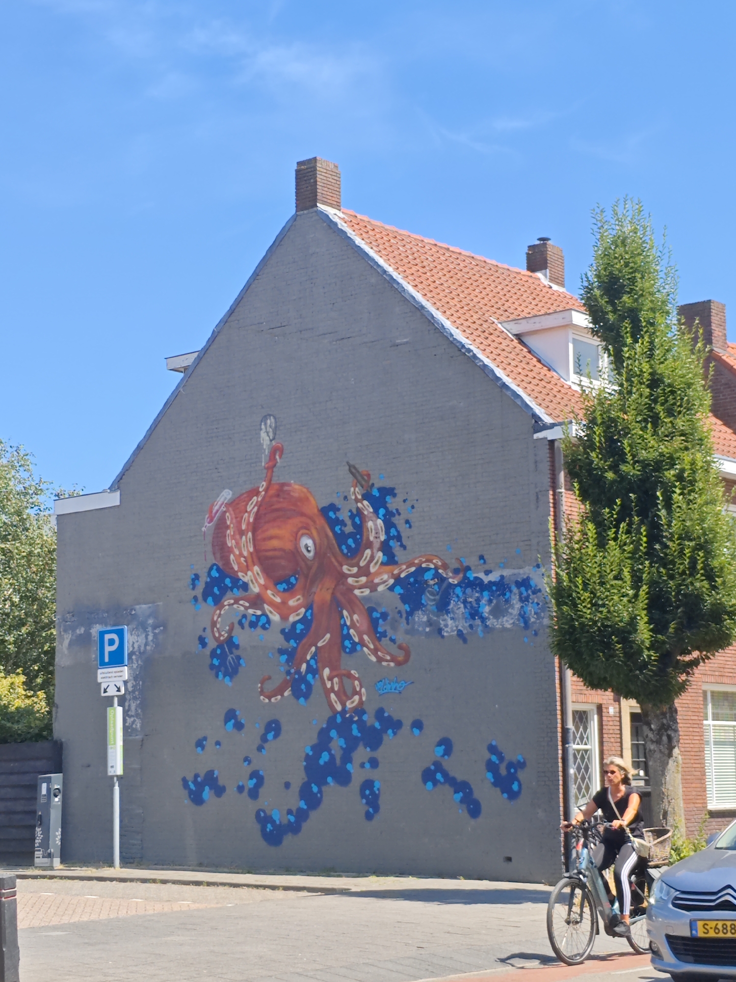 An outdoor, eye-level shot shows a grey brick building with an orange octopus mural painted on its side. A woman wearing sunglasses, a black top, and striped leggings is riding a bicycle with a basket on the front. A silver car is parked to the right of the woman. A tree with green leaves is positioned to the right of the building. A blue and white parking sign is visible on the left side of the building. The sky is clear and blue. The image appears to be taken in Tilburg, Netherlands.