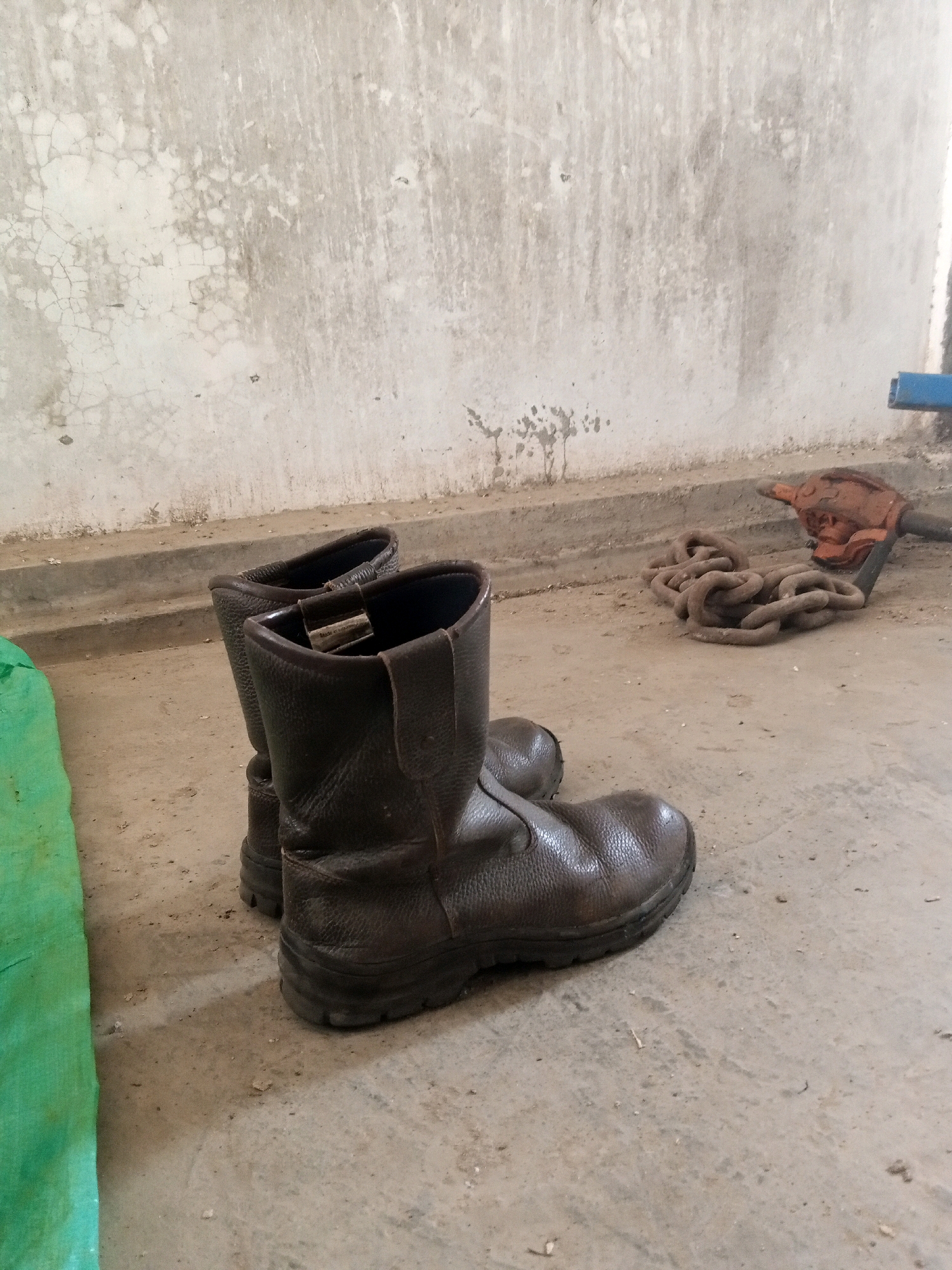 A pair of dark brown, worn leather work boots stand on a concrete floor. The boots have a thick sole and a reinforced toe. To the right of the boots, a thick metal chain is coiled on the ground, partially covered by a rusty orange tool that appears to be a jack or hoist. In the background, a rough, textured concrete wall shows signs of age and wear, with patches of discoloration and cracking. To the left of the boots, a portion of a bright green tarp is visible. The scene appears to be an outdoor or partially enclosed industrial or storage area, possibly located in Teluk Dalem Ilir, Indonesia, given the contextual information provided. There are no people visible in the image.