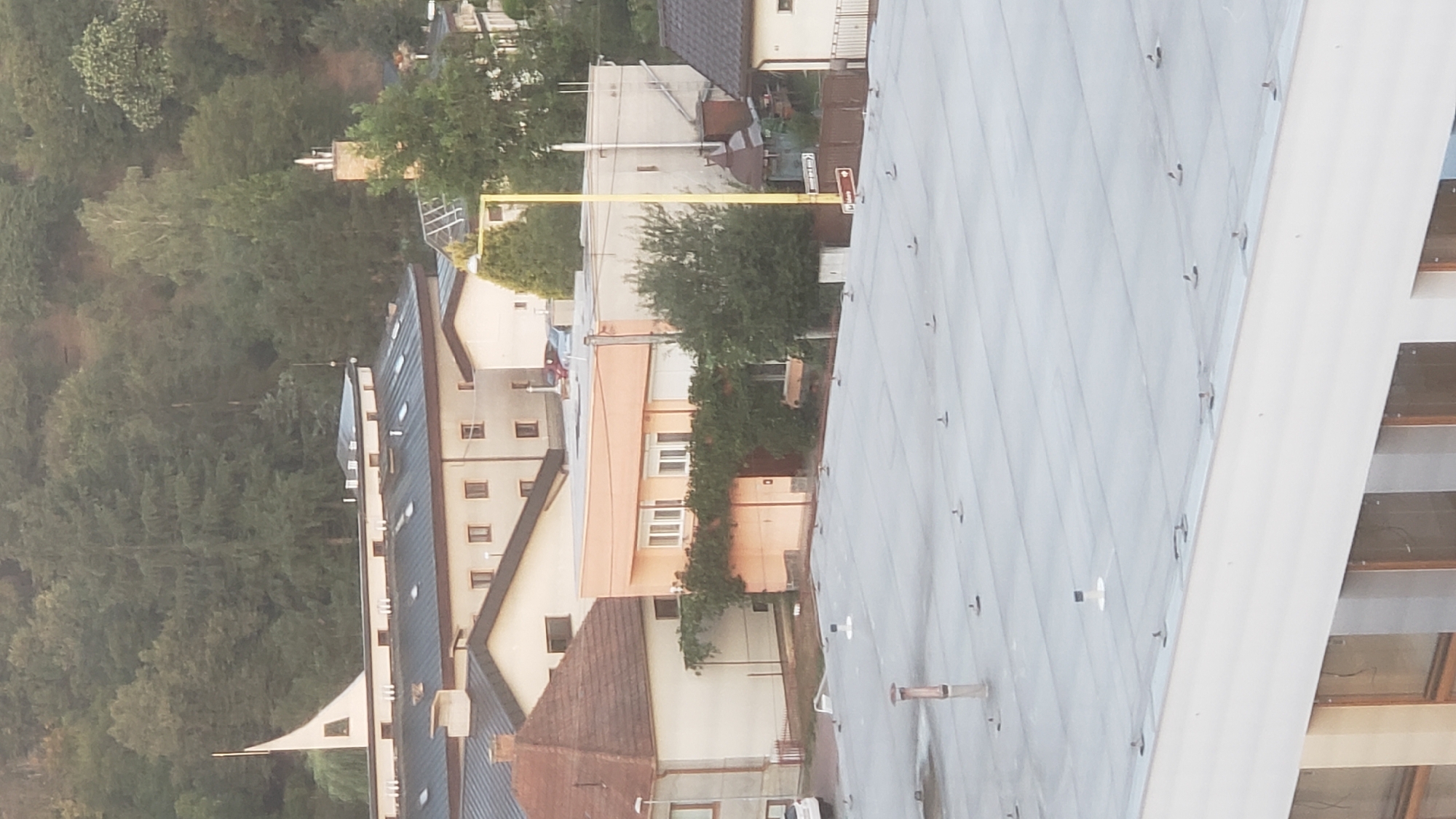 The image shows an elevated view of a town with several buildings and dense trees in the background. In the foreground, a light gray, flat rooftop with numerous small anchors or fixtures is visible. Moving further back, there are several multi-story buildings with dark roofs and numerous windows. Some buildings have beige or light orange facades. One building has a distinct curved roofline and a white spire-like structure on top. A bright yellow lamppost stands near a building with a light beige facade. In the mid-ground, residential houses with terracotta tiled roofs are visible, with some covered in ivy. A white car is parked on the left side of the image. The background is dominated by a steep hill covered in lush green trees. There are no people or animals visible in the image. The scene appears to be set during daylight with the sun likely low in the sky, creating soft lighting. This view is consistent with an area within the city of Prešov, Slovakia, potentially an elevated residential or mixed-use district bordering a forested area.