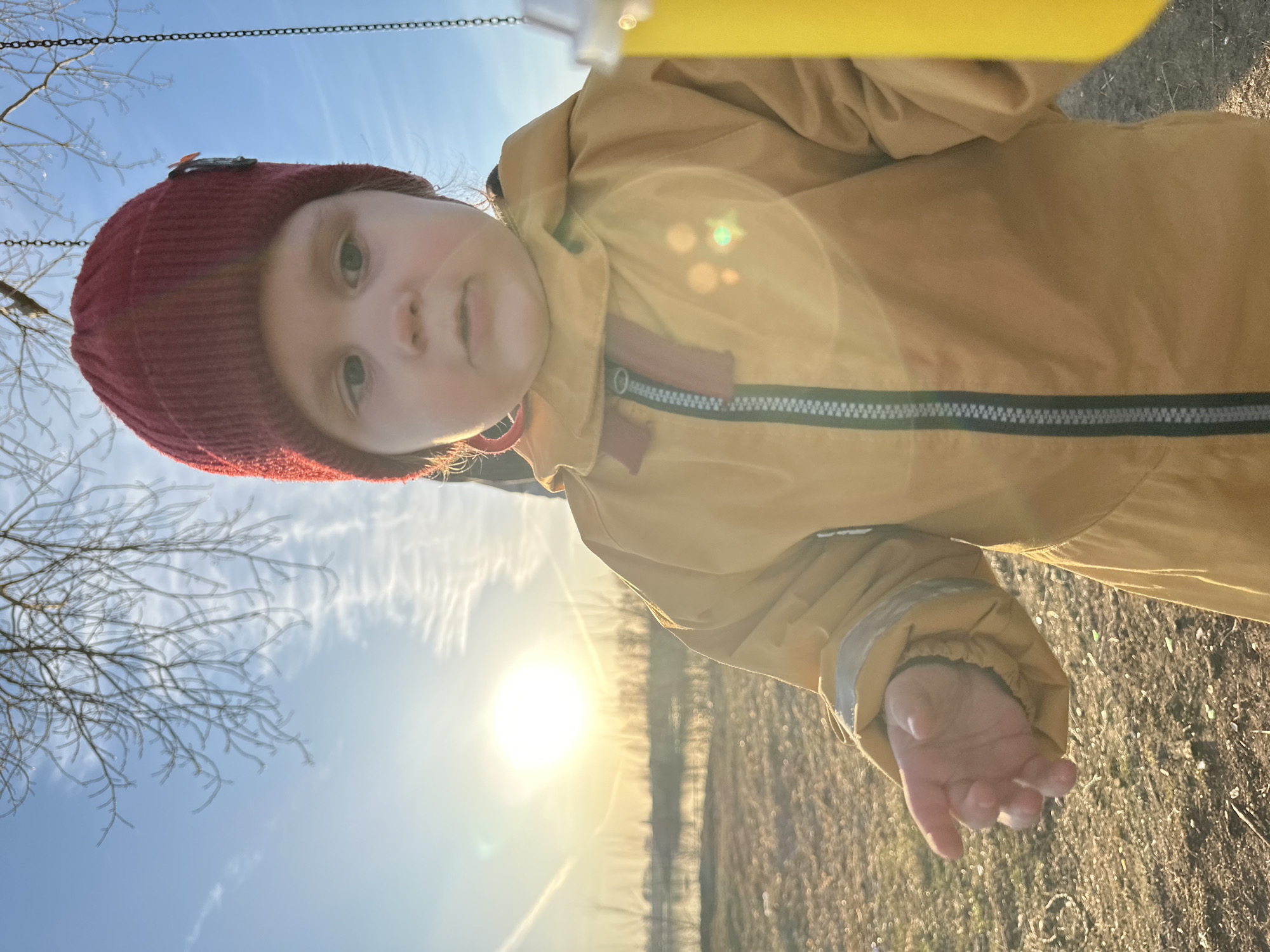 The image is presented with a vertical orientation, tilted counter-clockwise, showing a young child from a low-angle perspective outdoors. The child's face, partially obscured by a bright sun flare, looks towards the viewer. They are wearing a reddish-brown knitted hat and a mustard-yellow or light-brown waterproof jacket or suit, featuring a dark grey zipper and a reflective strip. The child's left hand is visible at the bottom right, slightly open.
Above the child's head, chains of what appears to be a swing set are visible against a bright blue sky with wispy clouds. Bare tree branches, possibly dusted with frost or ice, extend into the scene from the left. A bright yellow object, likely part of a swing seat, is seen at the top of the frame. The lower half of the image, to the right of the child, is dominated by the setting or rising sun, which casts a warm glow and creates a significant lens flare. The ground beneath the child is covered in dry grass or sparse vegetation.
The presence of the sun low in the sky, combined with the bare, possibly frosted trees, suggests a cold season, such as winter or early spring/late autumn, and either early morning or late afternoon/evening. The child appears to be playing, likely on a swing in an outdoor setting, such as a park or a rural area. Given the provided context of Chaikovsky, Russia, the cold weather cues are consistent. No discernible text is visible in the media file.