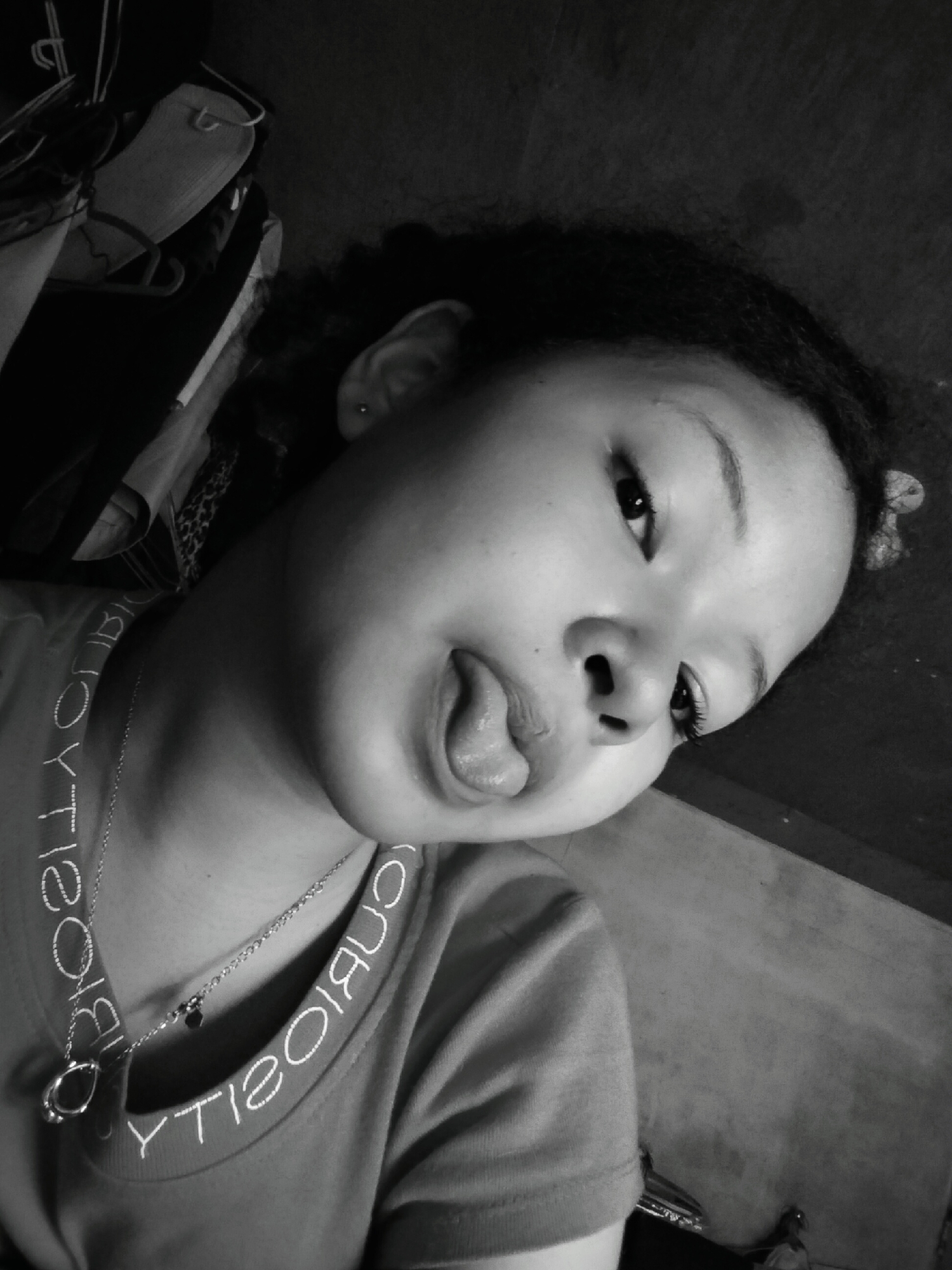 A young person with dark, curly hair is shown in a close-up, black and white, tilted shot. The person has their tongue sticking out slightly, with their mouth open. They are wearing a t-shirt with white lettering around the collar and a silver necklace with a pendant. In the background, there is a glimpse of clothing on hangers, suggesting a closet or wardrobe area. The setting appears to be indoors, possibly a residential space. The image does not provide any specific visual cues to pinpoint a location within Davao City, Philippines.