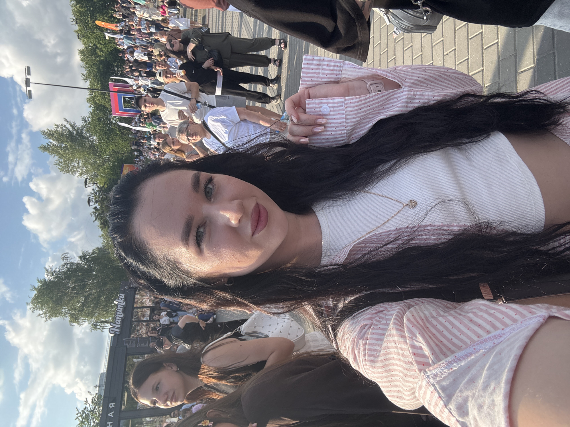 A woman is taking a selfie outdoors in what appears to be a public square or event space. She has long, dark hair and is wearing a pink and white striped shirt over a white top and a gold necklace. She is looking directly at the camera and smiling. In the background, a crowd of people is visible, many of them standing and looking around. Some are holding drinks, and some appear to be talking to each other. There are trees and buildings in the distance, as well as a cloudy sky. The ground is paved with interlocking stones. Based on the signage that says "Hanumku" and "НАС", the location is likely a street or public space in Novosibirsk, Russia.