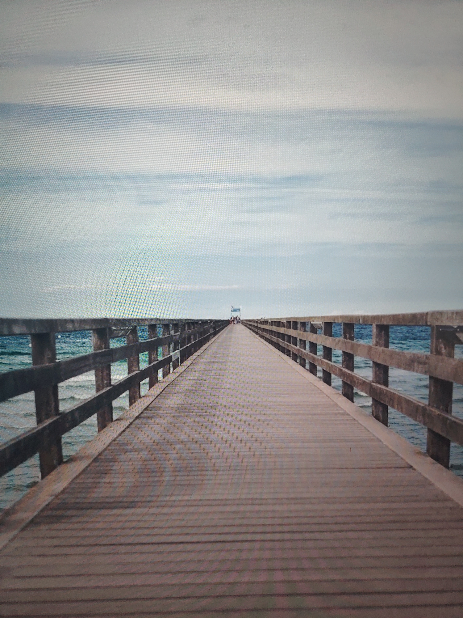 The image depicts a long, wooden pier extending into a body of water, likely the sea, under a cloudy sky. The pier is lined with wooden railings on both sides, creating a path towards a structure or platform visible in the distance. The water appears choppy, with visible waves. In the distance, there are figures of people. The location is likely coastal. The city and region are unknown.