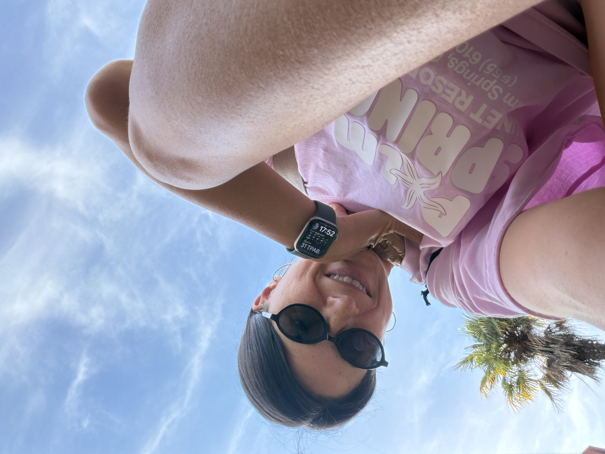 The image presents a low-angle, upward-facing view of a person wearing sunglasses and an Apple Watch, seemingly overlooking the camera. The watch displays a time of 17:52 and the date 31 ТРАВ. The person is wearing a light pink T-shirt with the word "PRIN" in white block letters, along with "NET RESO" and "m Springs, (555) 610" printed below. A palm tree is visible in the background. The sky is a light blue with white clouds. The location in the city of Budva, Montenegro is not discernible from the image.