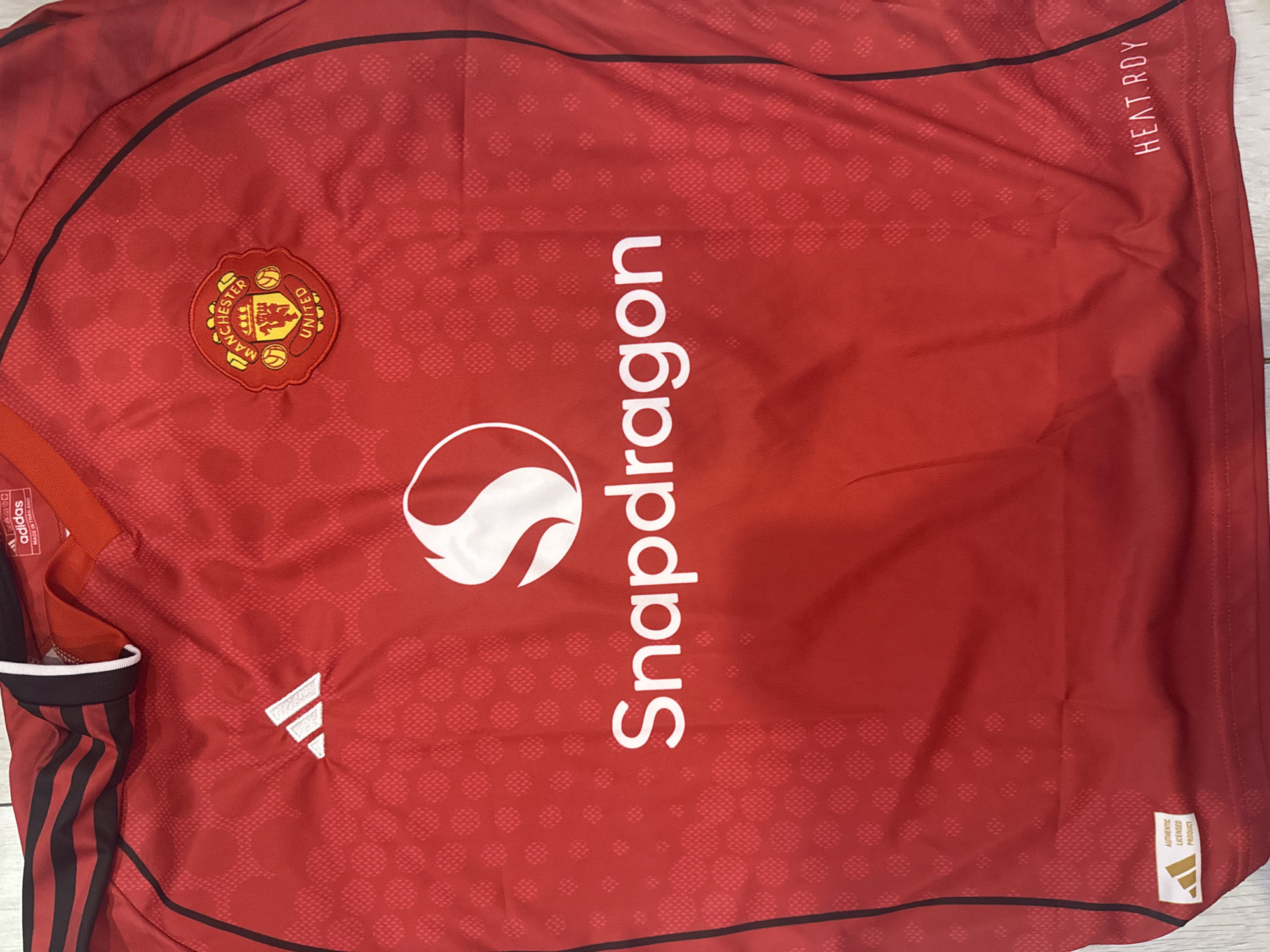 The image shows a close-up of a red Manchester United football jersey with black stripes on the shoulders. The jersey features the Manchester United crest on the left chest, an Adidas logo on the right chest, and a large white Snapdragon logo in the center. An "Authentic Licensed Product" tag with an Adidas logo is visible on the bottom left hem, and the "HEAT.RDY" logo is on the bottom right hem. The jersey is laid flat on a light-colored surface, possibly a table or floor, with some visible folds and wrinkles indicating it is a fabric item. There are no people, actions, interactions, or any elements that would suggest a location in Burgas, Bulgaria. The image is a product photograph of apparel.