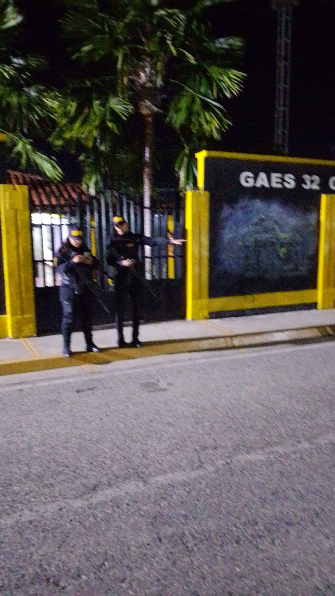 The image captures a nighttime scene in Tinaquillo, Venezuela, featuring two security personnel standing near a gate and a wall. The setting appears to be an official or guarded entrance, illuminated by artificial light against a dark sky.
Two individuals, likely security guards, are positioned on a sidewalk in front of a dark, wrought-iron gate and a yellow-framed wall. Both are dressed in dark uniforms, including long-sleeved shirts or jackets and pants, and wear yellow-brimmed caps. The person on the left is looking down, seemingly at a mobile phone held in their hands. The person on the right is standing with their right arm extended slightly, pointing or gesturing towards the right side of the frame, and appears to be holding a long, thin object, possibly a baton.
Behind the personnel, a tall, dark gate is visible, with a building structure featuring horizontal slats or windows beyond it. To the right of the gate, a large dark wall is framed by bright yellow vertical and horizontal beams. White text on this wall clearly reads "GAES 32 C". The letters "GAES" likely refer to the "Grupo Antiextorsión y Secuestro" (Anti-Extortion and Kidnapping Group) of the Venezuelan National Guard. Above the gate and wall, several large palm trees are prominent against the dark background, their fronds green even in the low light. In the foreground, a dark asphalt road is visible. The overall mood suggests a security presence at a guarded location.