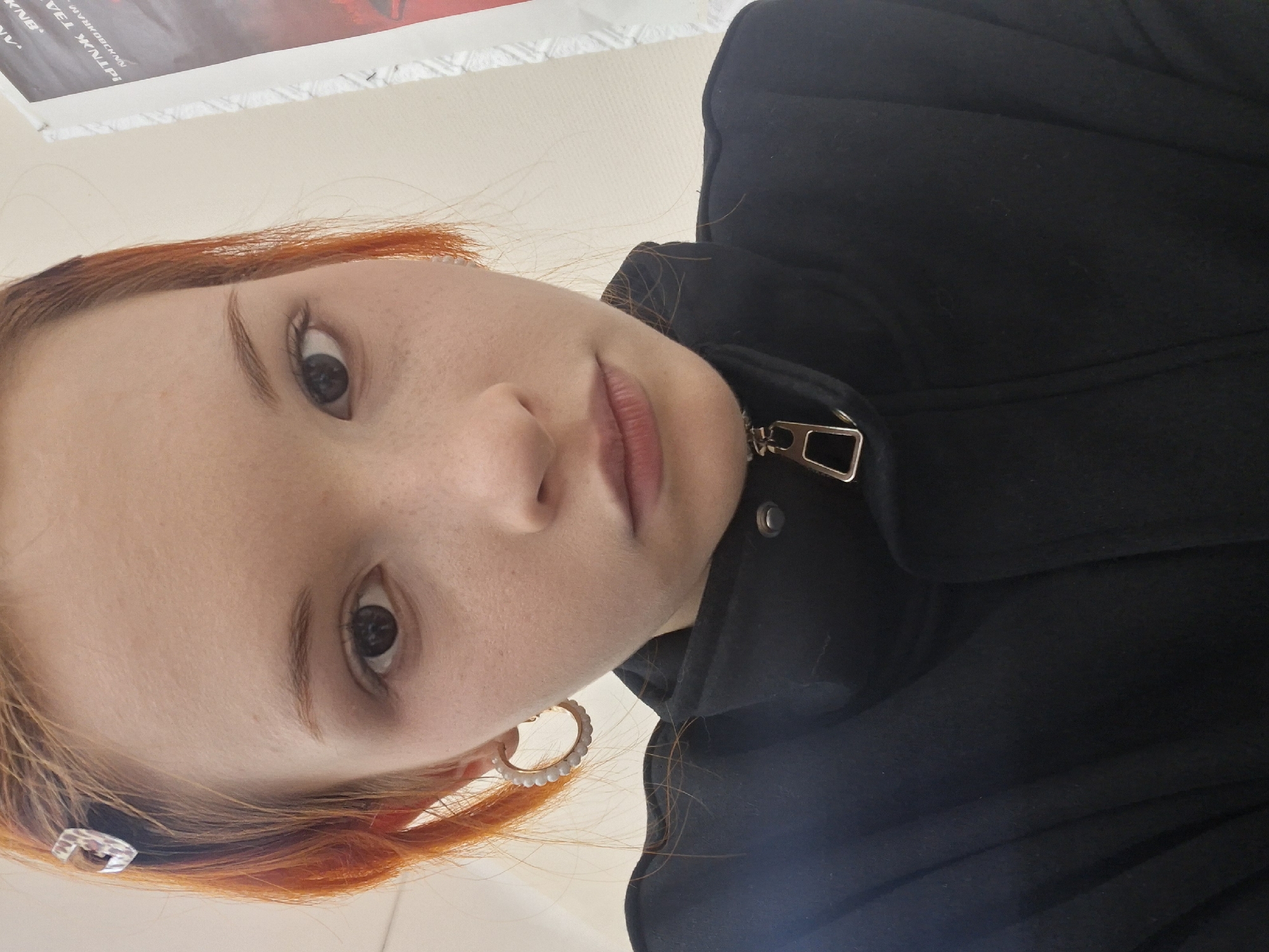 This is a close-up, upward-angled portrait of a young person, likely a selfie, taken indoors in Chaikovsky, Russia. The subject has reddish-orange hair, dark eyes, and visible freckles across their nose and cheeks. They are wearing a black coat or jacket, zipped up, with a prominent metallic zipper pull. A white, beaded hoop earring adorns their right ear, and a small clear hair clip is present in their hair on the left side of the frame. The person is looking directly at the camera with a neutral expression. The background features a light-colored wall, and in the upper portion of the image, part of a poster or artwork is visible. Text on this poster includes fragments such as "KNB", "VEL WULF", "METAUX TP", and "AN^". The lighting suggests it is daytime.