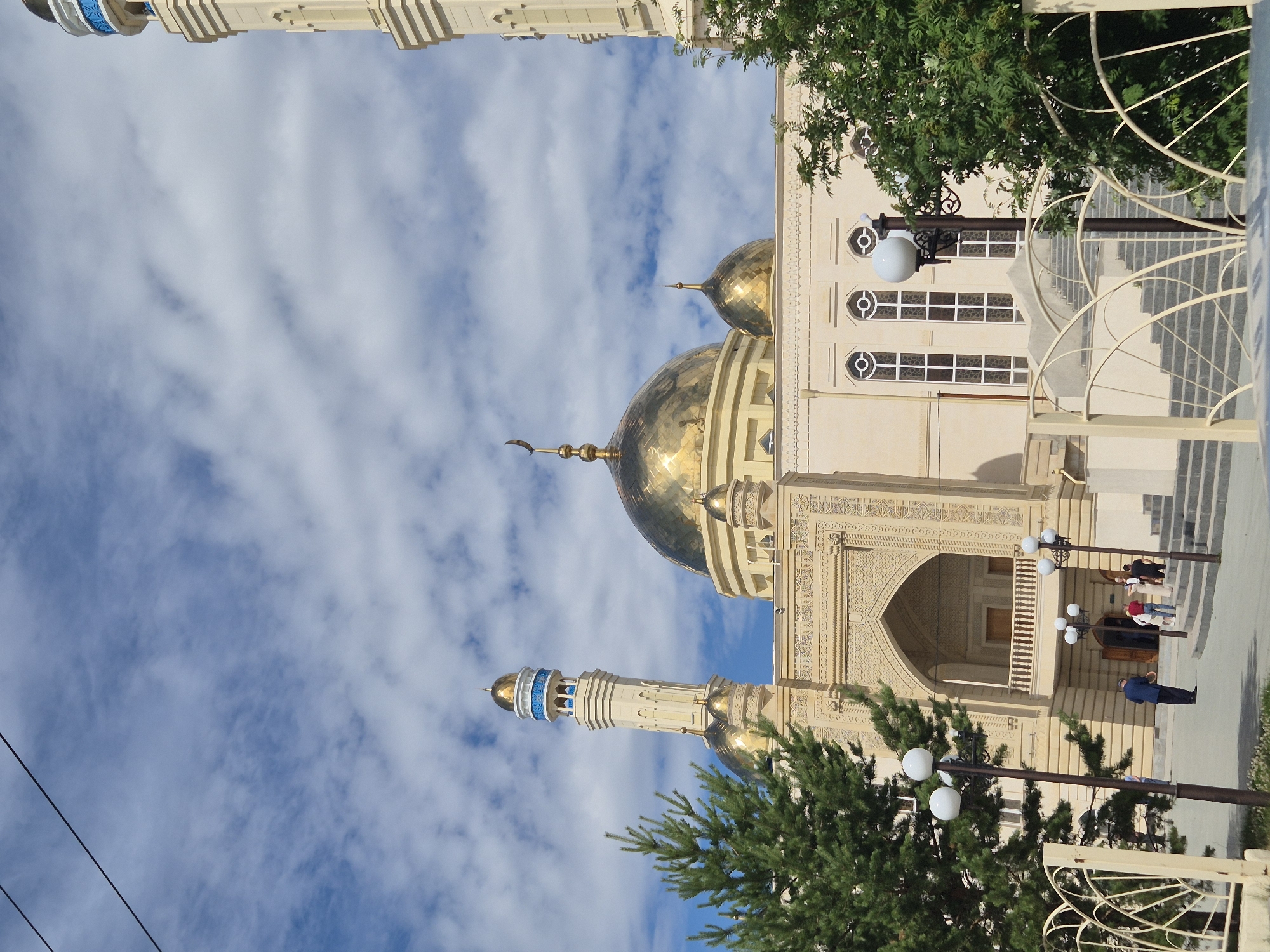 The image shows a large, ornate mosque with golden domes and intricate architectural details, set against a bright blue sky with scattered white clouds. The mosque features tall minarets, a prominent central dome, and decorative stonework. In the foreground, there are some people walking on a paved area and ascending a set of stairs leading to the mosque. Trees and streetlights are also visible. The scene appears to be in an urban setting, likely a public space in Kokshetau, Kazakhstan.