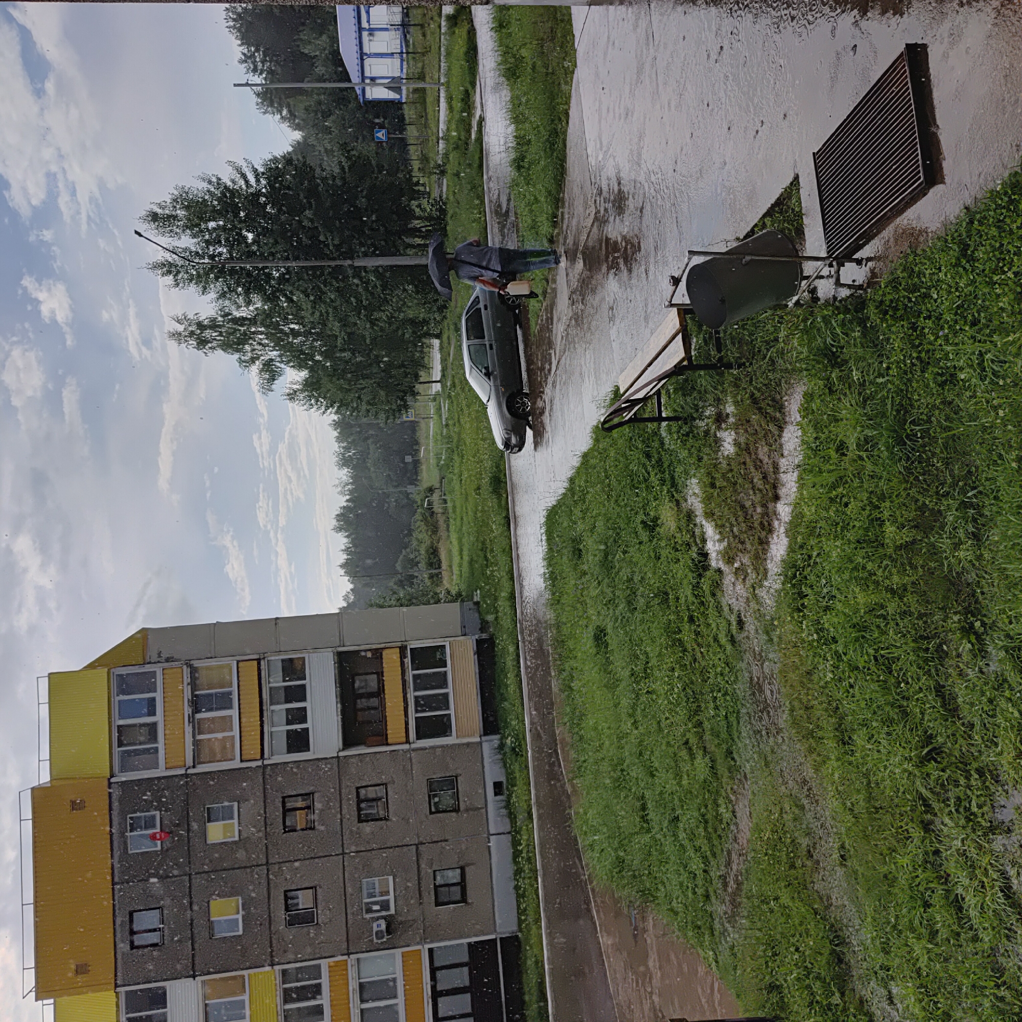 In the city of Markovsky, Russia, during a rainfall, a residential building with a yellow and gray facade stands on the left. In front of it, a paved area is flooded with water, reflecting the cloudy sky. A dark gray sedan is partially submerged in the water on the road. A person holding a black umbrella walks along a wet sidewalk, carrying a white jerrycan and a bag. A wooden bench and a trash can are situated on the grassy area next to the sidewalk. Further in the background, trees and other buildings are visible.