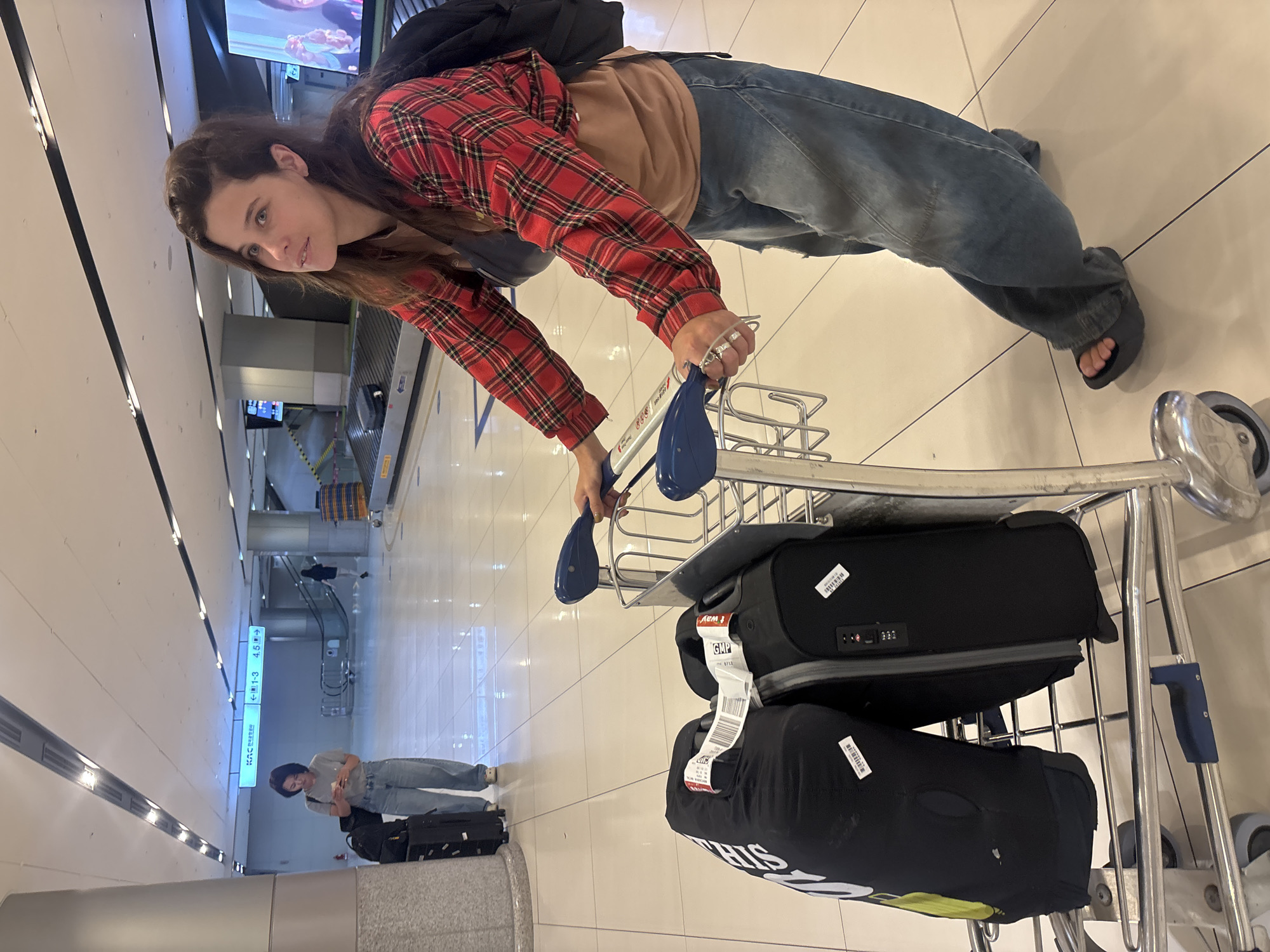This media file captures a woman pushing a luggage cart in an airport terminal. She is wearing a red and black plaid shirt over a light brown hoodie and blue jeans. Her hair is long and brown, and she is looking towards the camera with a slight smile. She is wearing silver rings on her right hand, which is on the handle of the luggage cart. The cart contains at least two black suitcases, one with a combination lock.
The scene is an indoor airport environment with a polished tile floor and white walls. Fluorescent lighting illuminates the space. In the background, there are signs and escalators, suggesting a busy transportation hub. One sign is partially visible with Korean text and numbers "1-3" and "4.5" with arrows, indicating directions or gate numbers. Another sign reads "KAC". A second person, wearing a grey t-shirt and blue jeans, is visible in the distance, also with luggage. The overall impression is one of travel and transit. The image is oriented vertically, giving a sense of looking down on the scene. The time of day cannot be determined from the image.