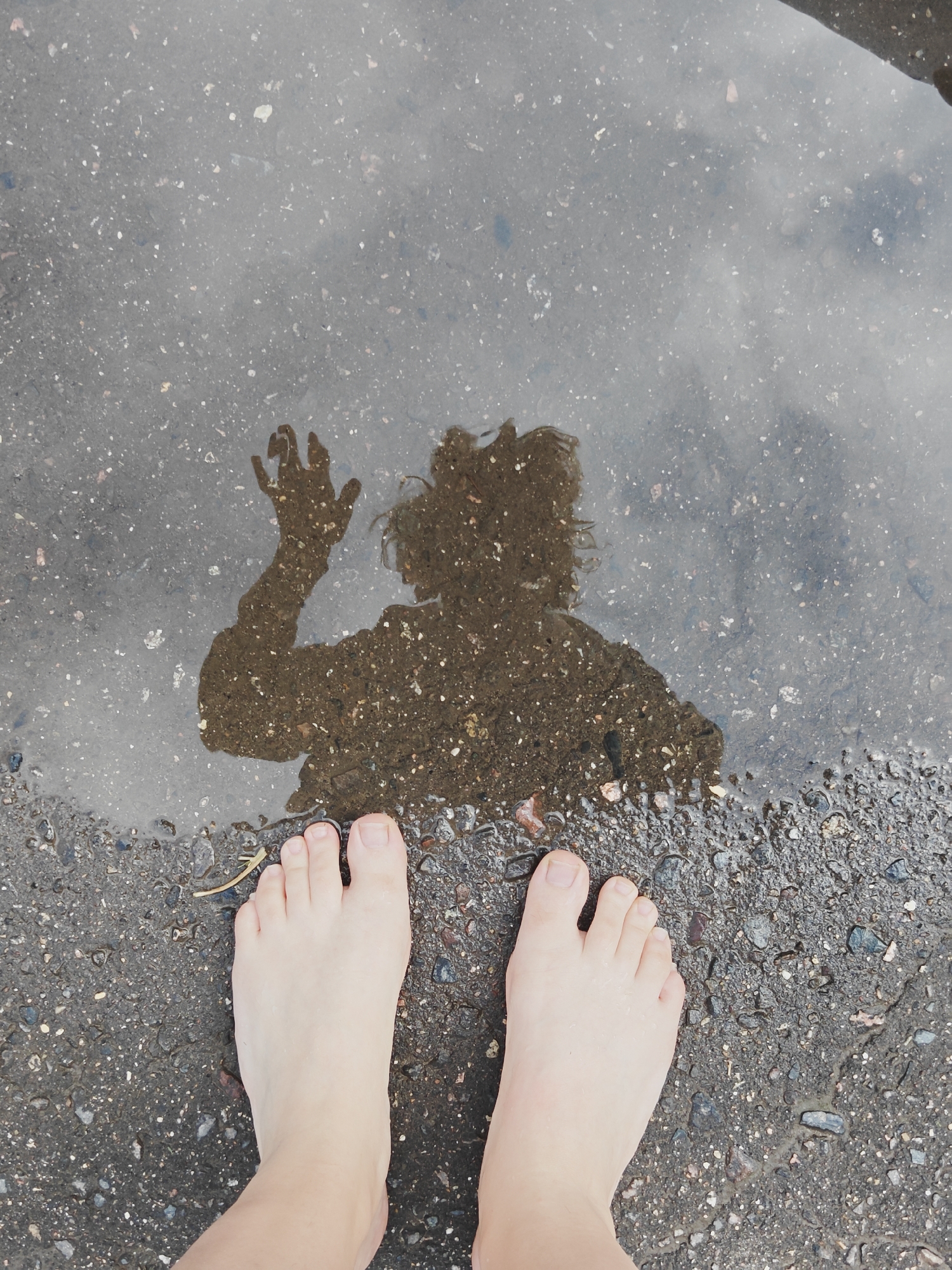 A reflection of a person is visible in a puddle on an asphalt surface, with the person's bare feet in the foreground. The reflection shows the person's head and raised right hand. The puddle is on a dark, wet asphalt ground with small stones and debris scattered on it. The scene appears to be in a public area, possibly a street or sidewalk, and the lighting suggests it is daytime. The location is identified as Koledino, Russia.