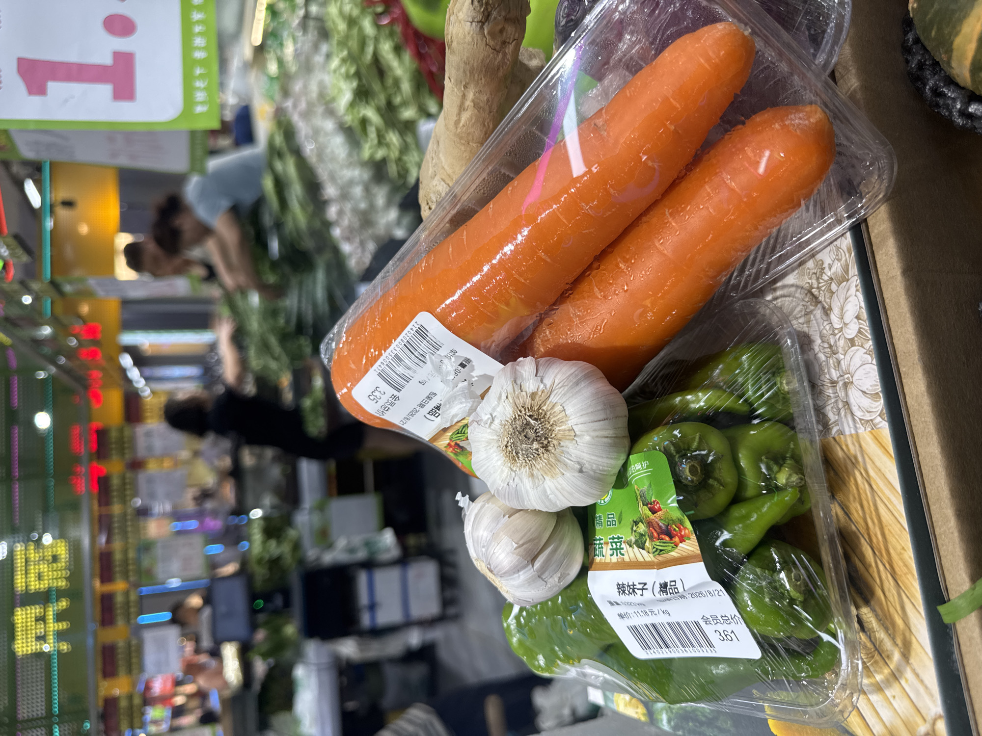 The image depicts a close-up view of a supermarket display of fresh produce, specifically in Shenyang City, China. In the foreground, a clear plastic container holds several vibrant green chili peppers. Adjacent to the peppers are two bulbs of garlic. To the right of these are two bright orange carrots, also enclosed in clear plastic packaging. The background is blurred, but it shows a supermarket environment with shoppers and other produce. A sign with the number "1" in large pink font is visible in the upper right corner, suggesting a price or section marker. There are also labels with Chinese text on the produce packaging, providing product information and possibly pricing. The overall impression is that of a well-stocked grocery store.
