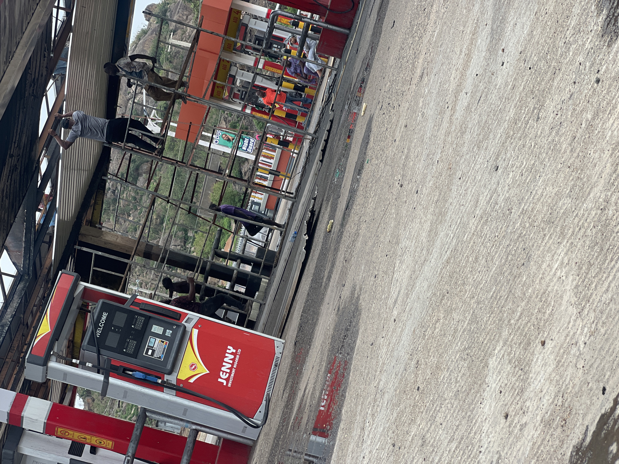 A street-level, tilted view reveals a Jenny Investment Nigeria Ltd. gas station located in Gura Top, Nigeria. The gas pump, predominately red with yellow accents, is labeled "WELCOME" on the display screen. Behind the pump, a metal structure supports an orange canopy overhead. Several individuals are visible, some standing and others climbing the metal structure. The background shows a hillside with sparse vegetation. The ground is concrete with visible wear and discoloration.