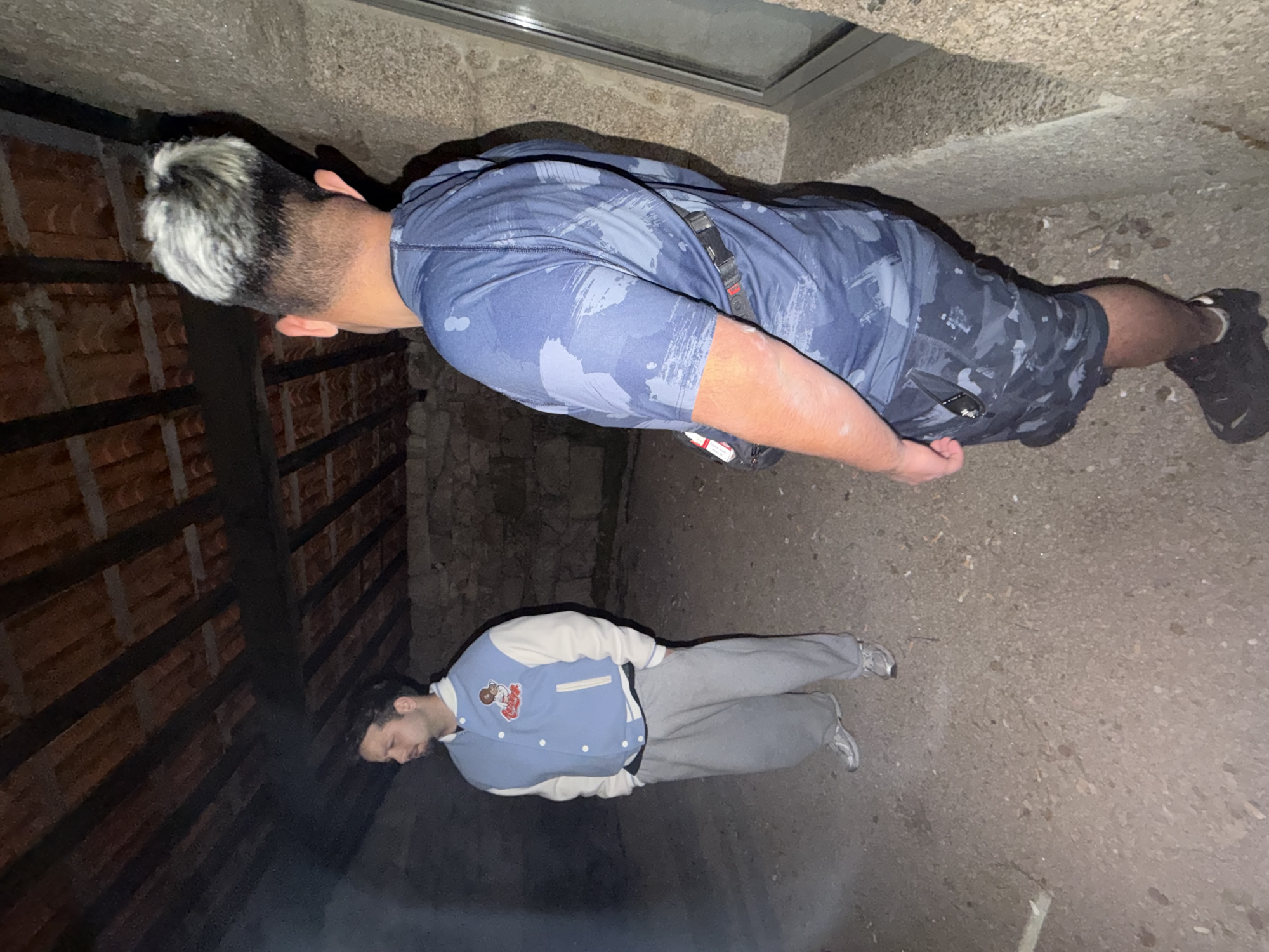 Two men stand in an outdoor location at night. The man on the top wears a blue t-shirt and shorts with dark shoes. The man on the bottom wears a blue and white jacket with gray pants and silver shoes. The ground is tan in color. The location is in Vila Real, Portugal.