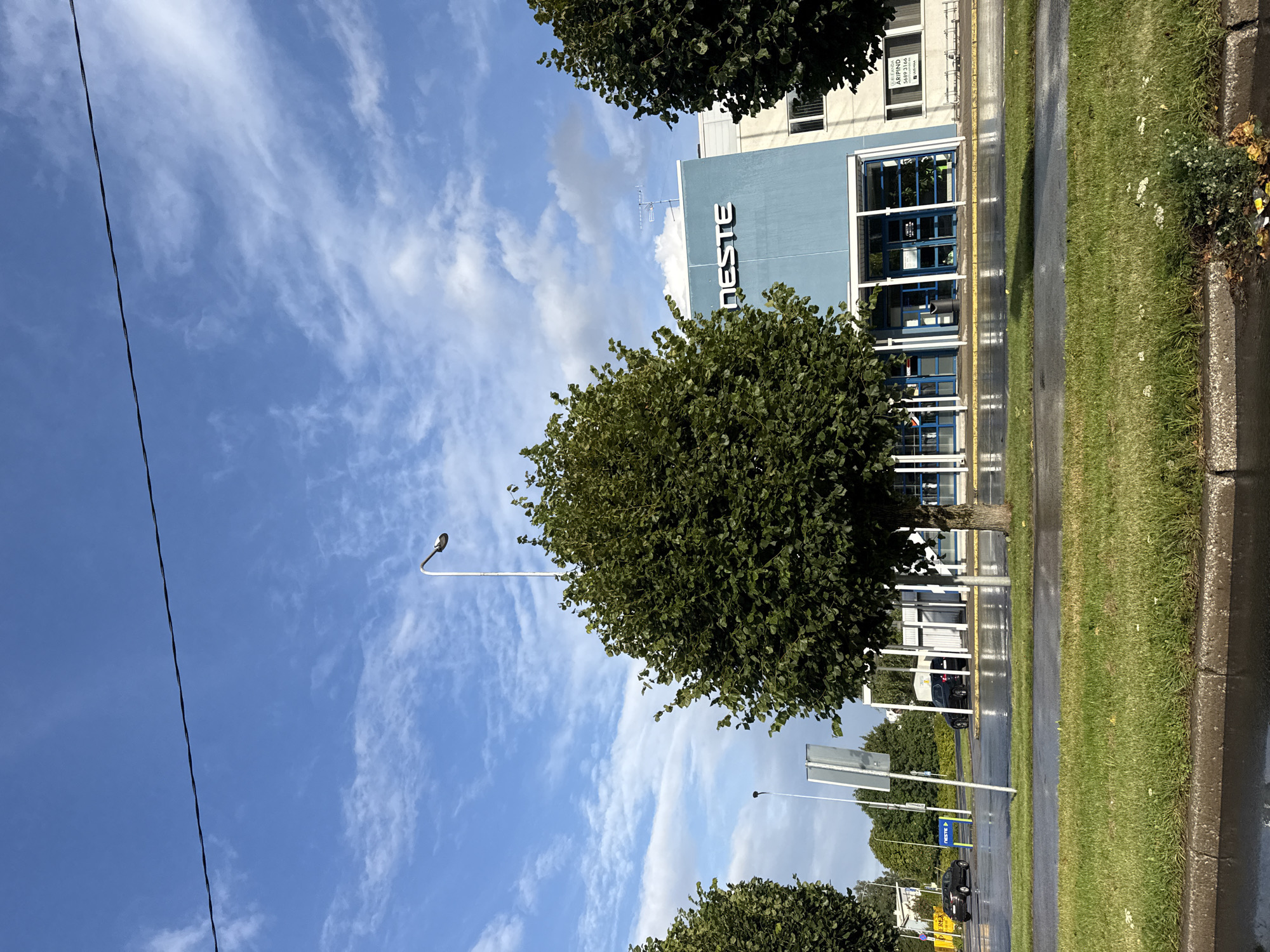 The image depicts a street view in Tallinn, Estonia, under a partly cloudy blue sky. In the foreground, a wet asphalt road runs horizontally across the frame, bordered by a strip of green grass and a concrete curb. Beyond the road, a modern building with a light blue facade and white accents is visible, displaying the white text "NESTE" prominently on its upper section. The building features a glass-covered entrance area with blue-framed doors. Trees with lush green foliage are positioned in front of the building, partially obscuring the view. A few vehicles are present on the road, including a dark-colored car. Streetlights and traffic signs are also visible in the background. The overall impression is that of an overcast day, possibly after a rain shower, with the wet road surface reflecting the sky and surroundings.