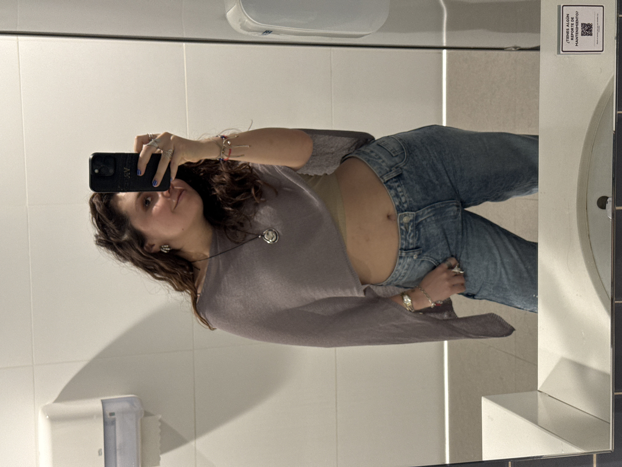 A woman is taking a selfie in a bathroom mirror. She is wearing a light purple top, blue jeans, a necklace, and multiple rings and bracelets. Her midriff is exposed. The bathroom has white tiled walls, a white sink, a white hand dryer, and a white paper towel dispenser. A sign with Spanish text and a QR code is affixed to the sink area. The location is unspecified but inferred to be in Mexico City, Mexico.