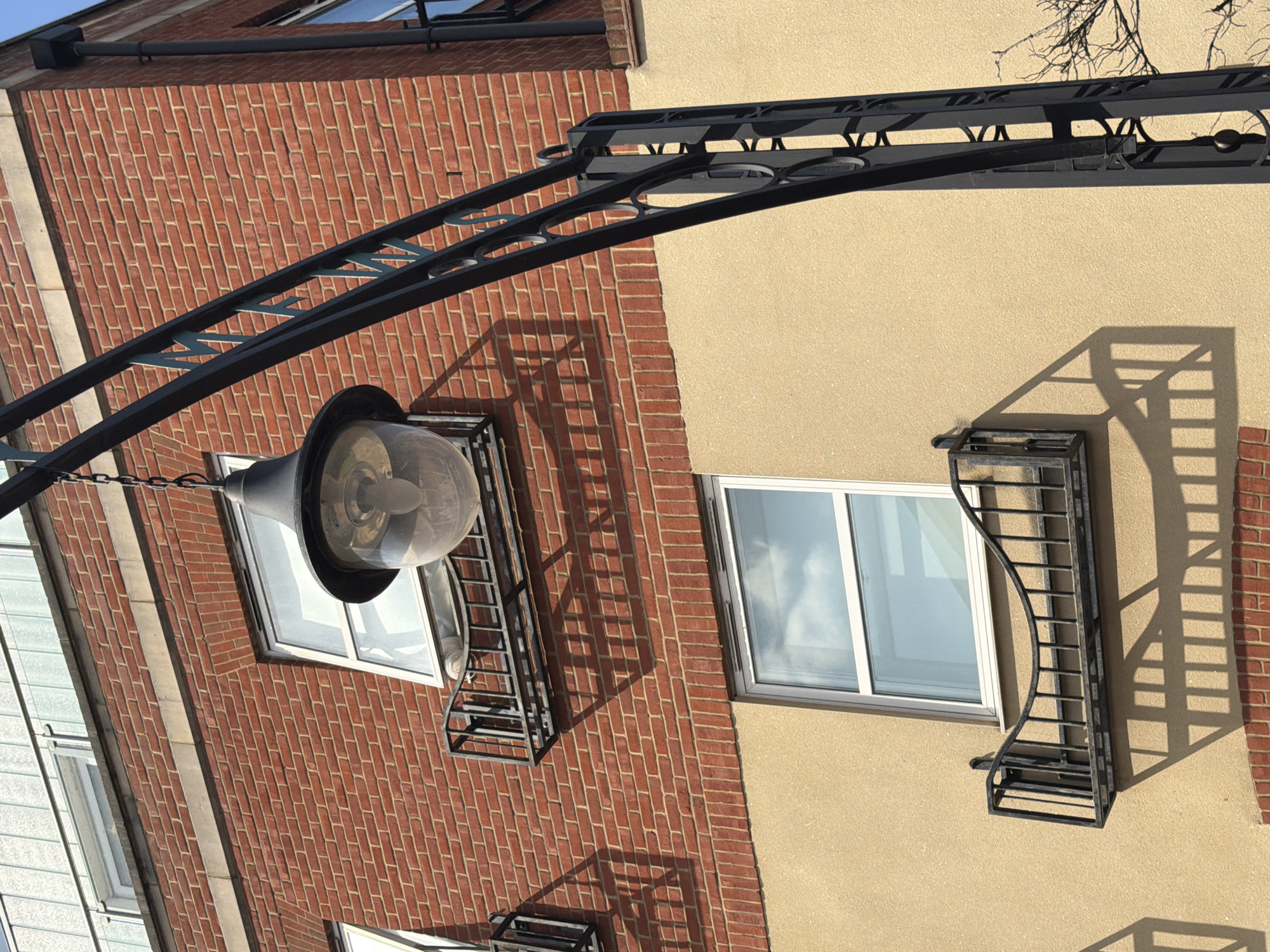 A street lamp hangs from a decorative black metal archway on the facade of a building in London, United Kingdom. The building features a red brick upper section and a beige stucco lower section. The upper section has two windows with small, decorative black metal railings in front of them. The lower section has one large window with a similar black metal railing. The street lamp itself is a clear glass orb with a black metal top and a visible lightbulb inside. Shadows of the lamp and railings are cast on the building's surfaces. A black downspout is visible on the right side of the brick section. A bare tree branch extends into the frame from the right.
