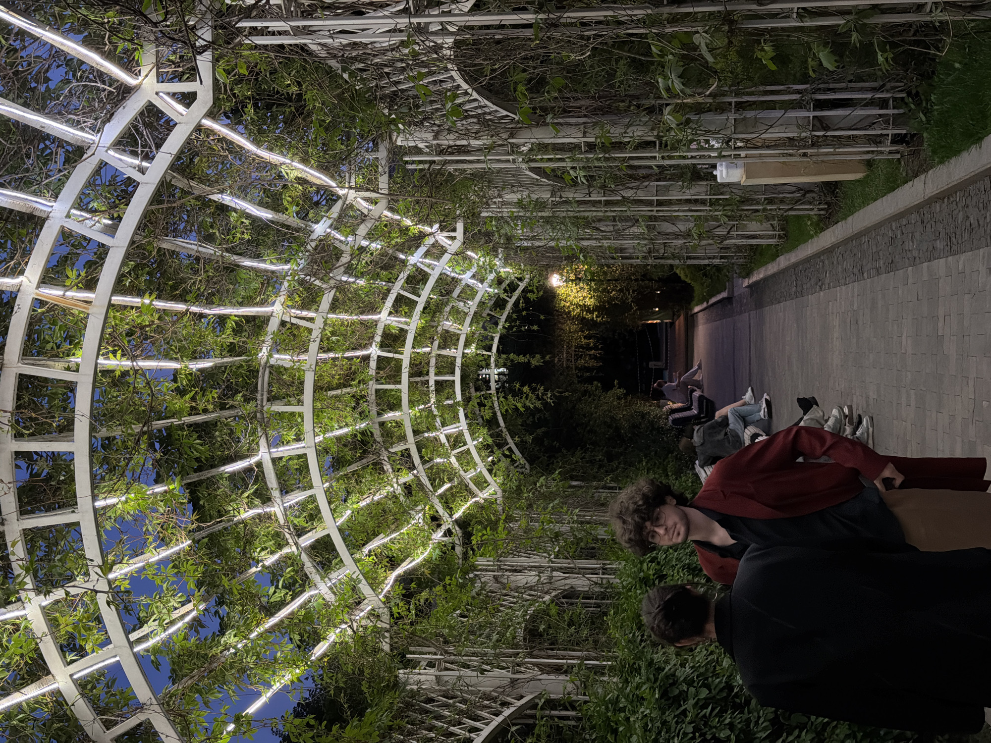 The image shows a nighttime scene in Moscow, Russia, within a pedestrian walkway or park area. A white arched trellis system covered in climbing vines stretches above a paved walkway. Light strips are installed along the trellis, illuminating the path. Several people are visible: two individuals are in the foreground, with one wearing a red shirt looking directly at the camera; the other person has their back turned. Further down the path, others are sitting or lounging on the pavement. The environment is characterized by greenery, including trees and shrubs lining the walkway. The location appears to be illuminated by artificial lighting.