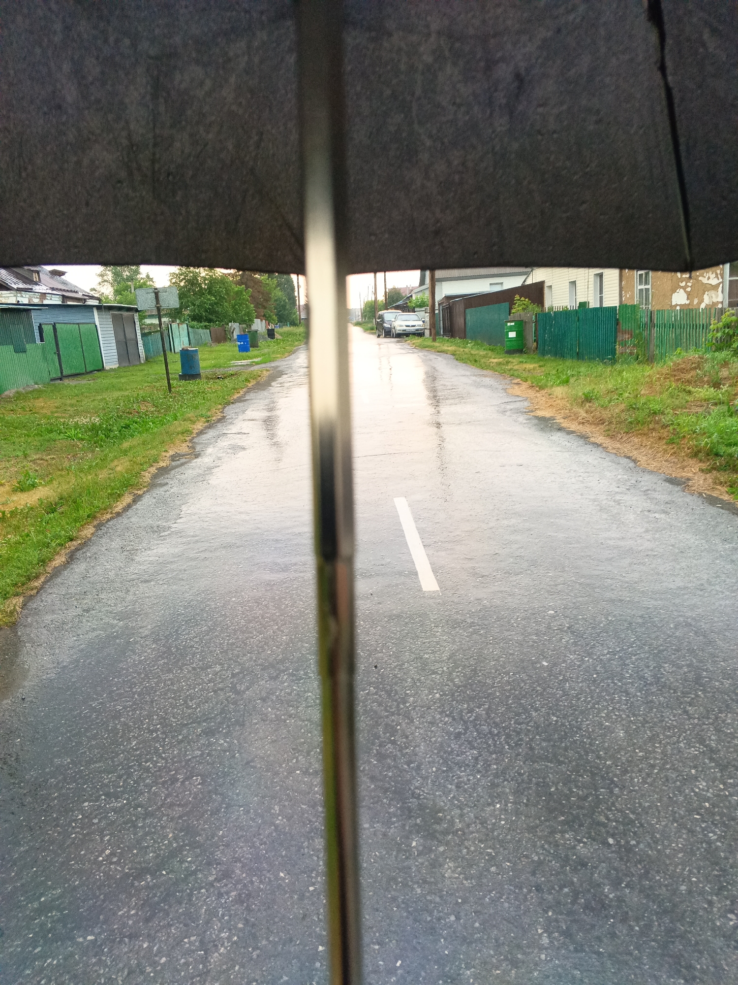 An eye-level, first-person perspective shot is taken from under a partially open, dark grey umbrella, looking down a wet asphalt road in a village setting. The road has a single white line down the center, indicating a lane. On the left side of the road, there are green wooden fences and outbuildings with green doors. Further down, there are houses and trees. On the right side, there is a green wooden fence and more houses, some with peeling paint. Two cars are parked on the left side of the road in the distance. The ground is wet, reflecting the sky and surroundings, suggesting it has recently rained or is currently raining. A metallic umbrella pole is visible in the center of the foreground, partially obscuring the view of the road. The general atmosphere is overcast. This scene is consistent with a rural or semi-rural area, possibly a village in Toguchin, Russia, given the architectural style and fencing.