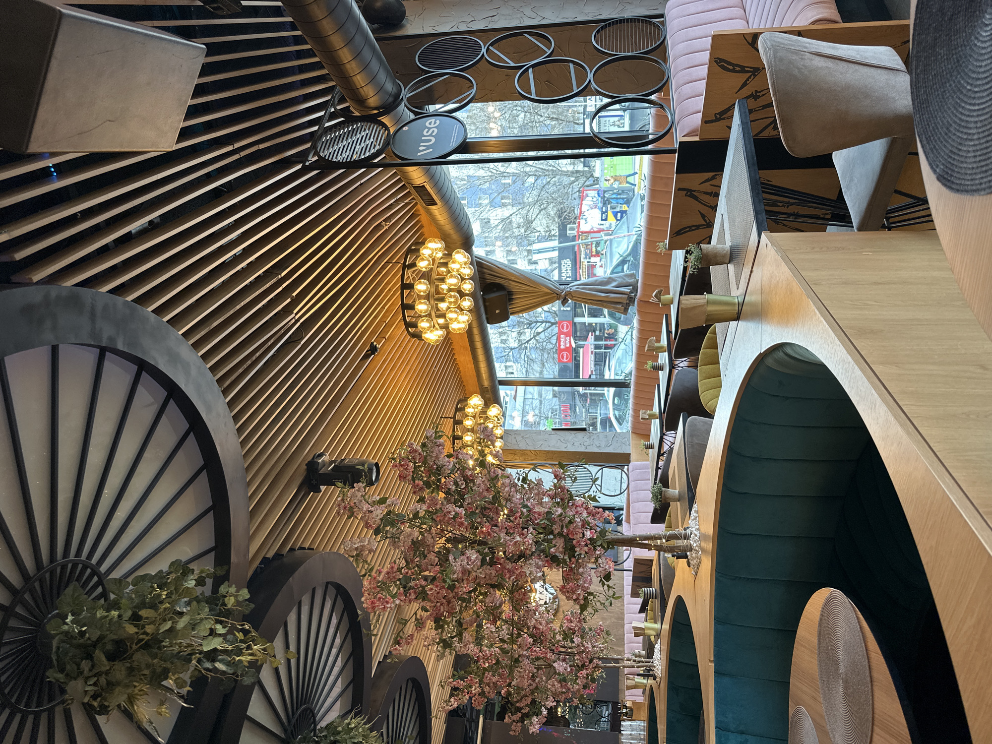 The image captures the interior of a restaurant, likely in Sofia, Bulgaria, judging by the 'V Use' sign visible through the window which aligns with the name of a shopping center in Sofia. The establishment is decorated with a mix of modern and rustic elements. Visible on the left side are large, decorative wagon wheels, foliage, and a slatted wooden wall. A faux cherry blossom tree with pink flowers is centrally placed. Ornate chandeliers hang from the ceiling. On the right, there are seating booths with teal interiors and wooden tables, complemented by pink upholstered banquettes. Through the window, the urban cityscape of Sofia is visible. The scene is taken from an elevated, slightly off-kilter angle. There are no people visible within the restaurant, but pedestrians and vehicles can be seen outside.