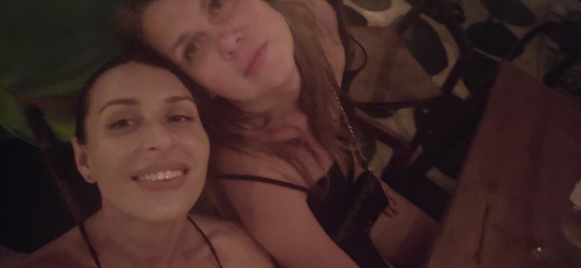 Two women are posing for a selfie in a dimly lit indoor environment. The woman on the left is smiling broadly, while the woman on the right is looking at the camera with a slight smile. Both are wearing spaghetti strap tops. The background is blurred and appears to be a bar or restaurant setting with a patterned wall covering and wooden furniture. There is a wooden table to the right. The location is Kuta Selatan, Indonesia.