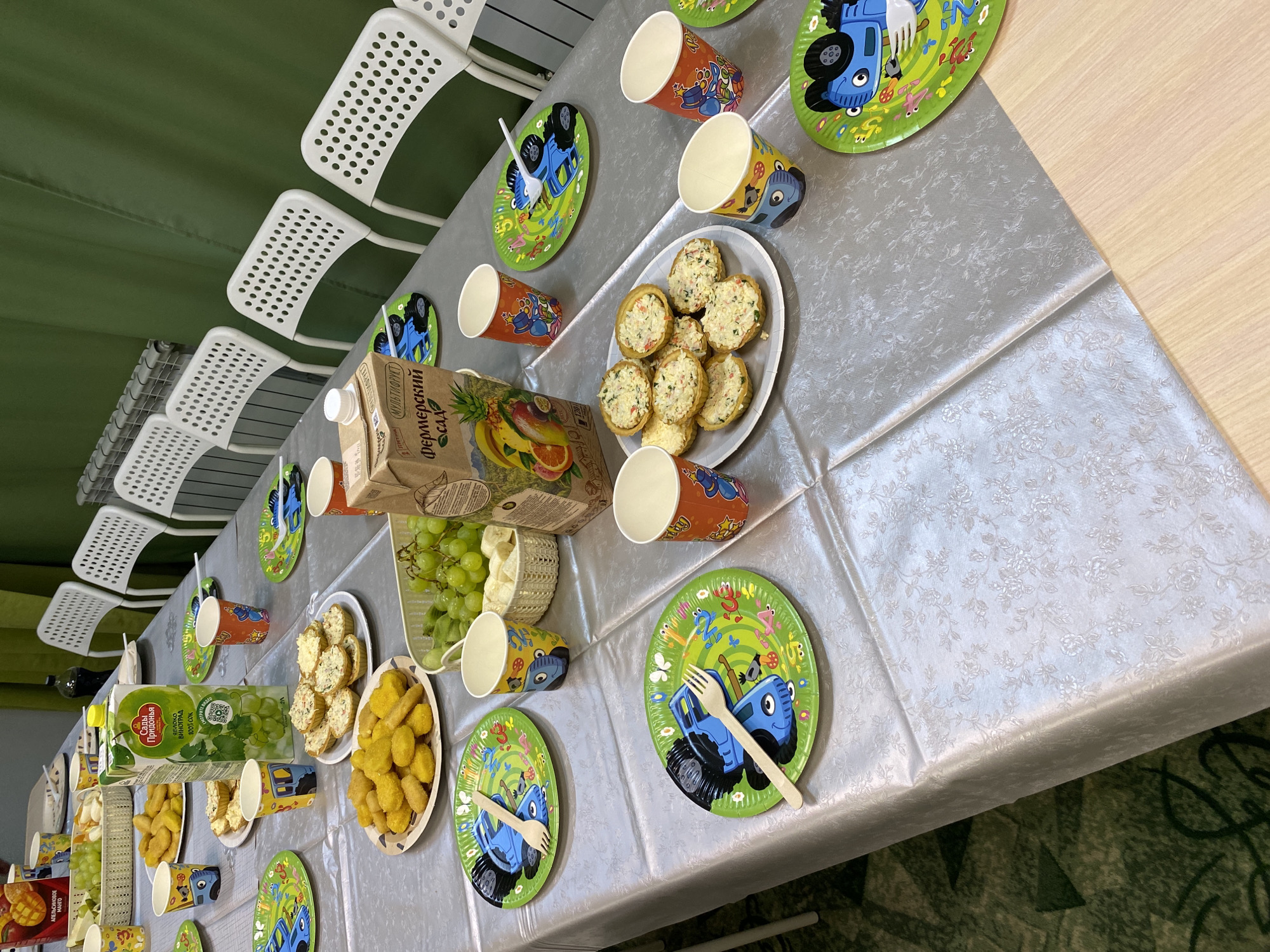 The image shows a children's party setup on a table covered with a light-colored floral patterned tablecloth. There are several white chairs lined up on one side of the table. The table is set with green plates decorated with cartoon tractors and wooden forks. There are also paper cups with cartoon designs scattered around the table. Plates of food, including what appears to be nuggets and filled tartlets, are present. A carton of "Sady Pridonia" apple-grape juice and another of "Fermerskiy Sad" multifruit juice are visible, along with a basket of green grapes. The location is Chaikovsky, Russia, as determined by the Cyrillic writing on the juice cartons and the name of one of the juice brands, "Sady Pridonia."