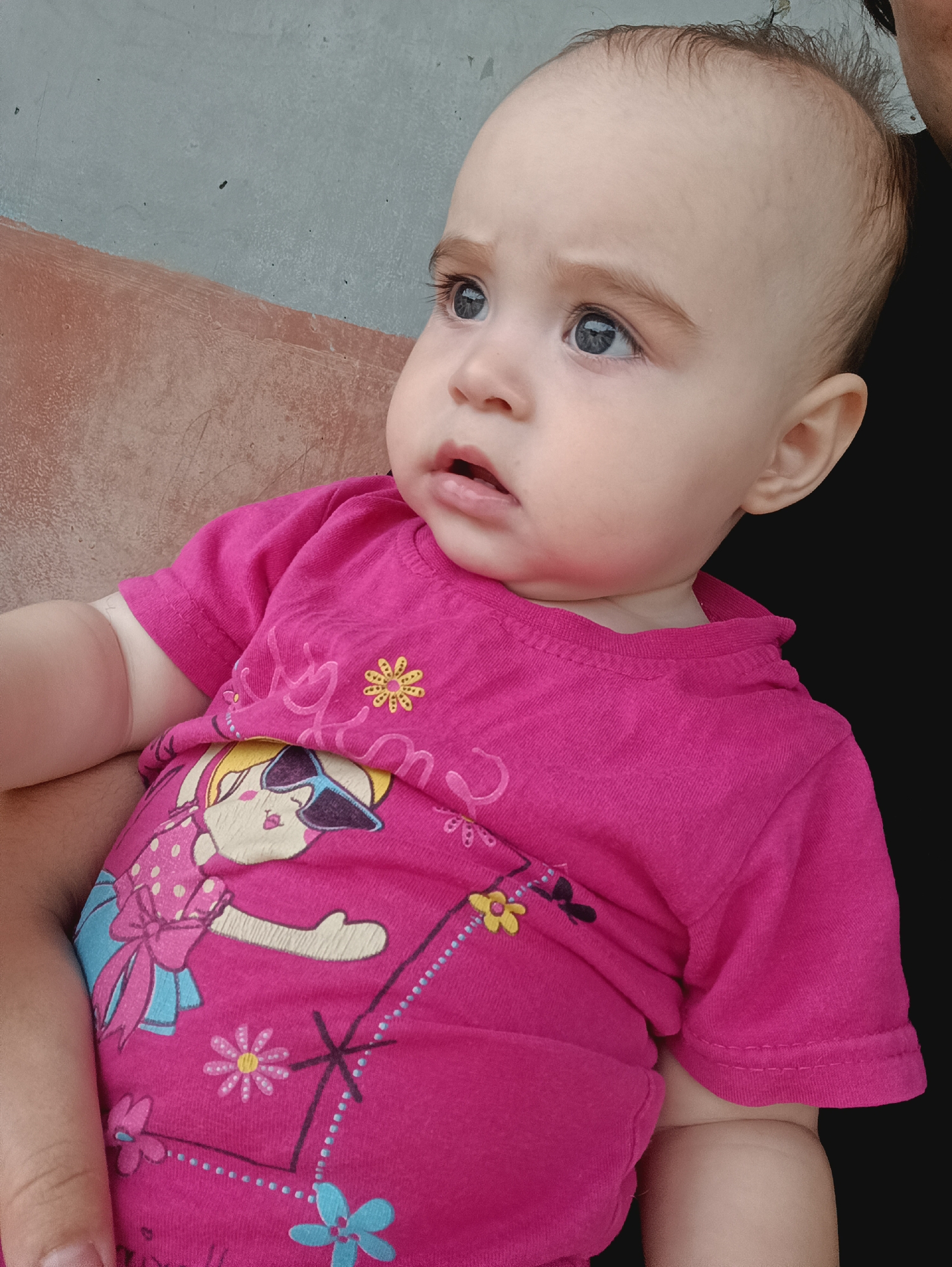 A young baby is held by an unseen person in front of a mottled wall that is half brown and half light blue, with a black background to the right. The baby, facing right with their head turned towards the viewer, wears a bright pink t-shirt adorned with cartoon graphics of a girl in sunglasses and floral patterns. The baby has large, wide-open blue eyes, light brown eyebrows and eyelashes, and sparse brown hair. Their mouth is slightly open, revealing pink lips. The lighting suggests an outdoor or well-lit indoor setting. There is no visible indication of the specific location within the city of Посад, Russia.