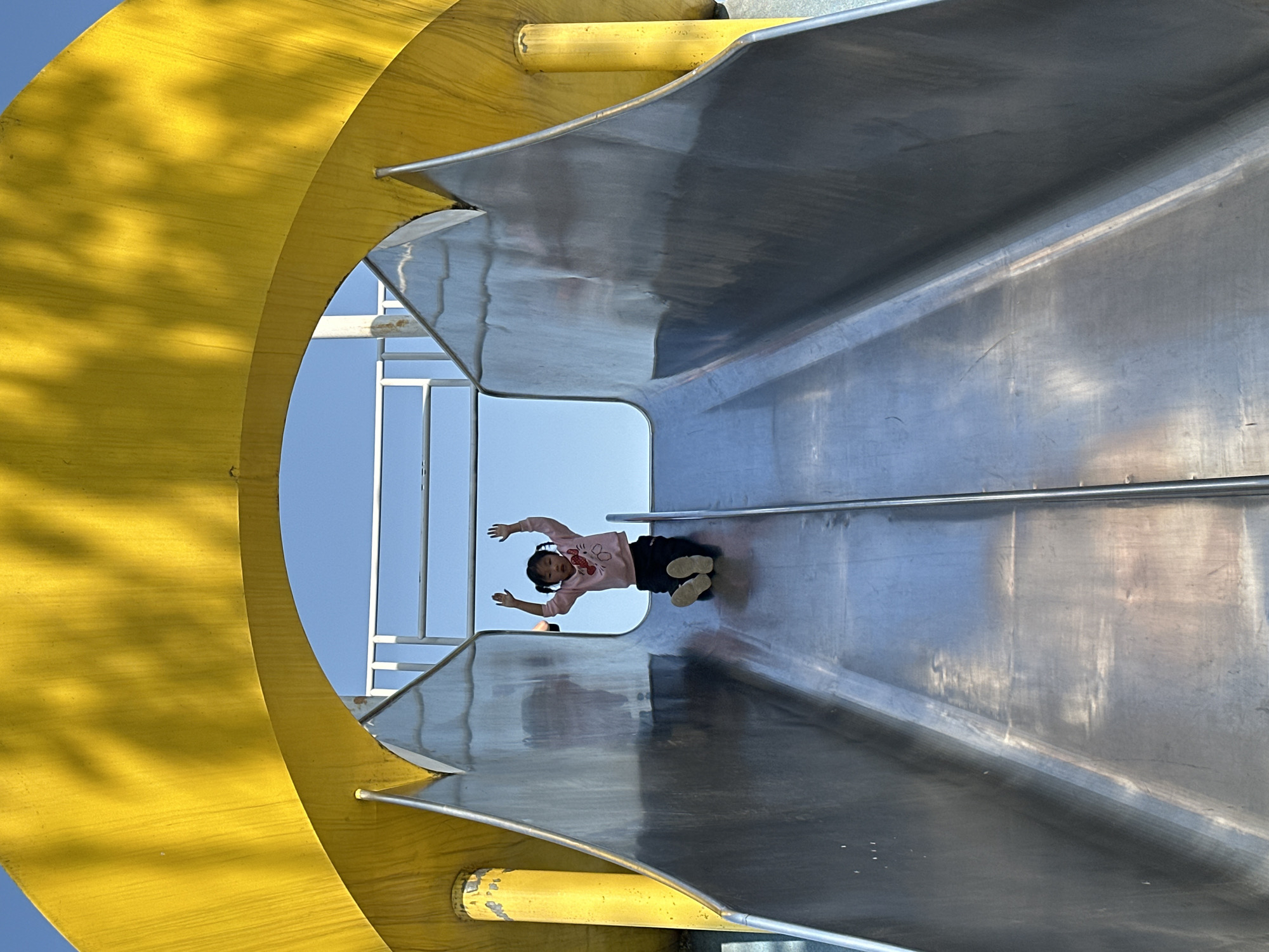 A low-angle shot depicts a child sliding down a wide, metallic slide at a playground. The slide is supported by a yellow, curved frame. The child, wearing a light pink long-sleeved shirt and dark pants, is positioned near the middle of the slide with arms outstretched. The surrounding environment shows a clear blue sky visible through the frame of the slide. The location is in the city of Jiangjin, China.