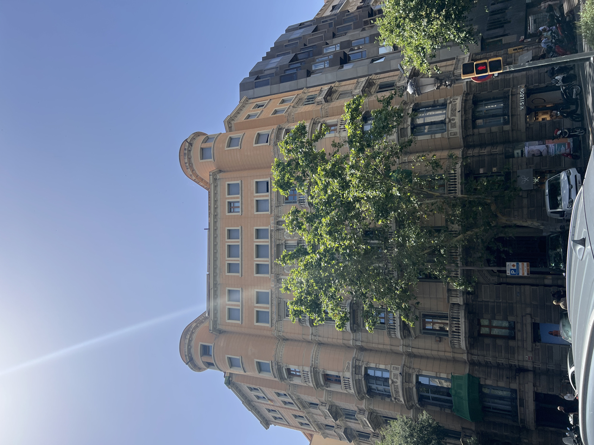 This media file shows an outdoor, upward-angled view of a multi-story building on a clear, sunny day, likely in Barcelona, Spain. The building exhibits a mix of architectural styles, with the lower floors featuring ornate detailing, curved facades, and numerous windows. Some of these lower sections are a pale peach color, while others are a darker stone. Above these, the building transitions to a more modern design with a stepped facade and numerous dark-framed windows. Lush green trees with dense foliage are visible in front of and between parts of the building.
The scene appears to be a city street, indicated by the presence of a traffic light with red and yellow signals, a "P" parking sign, parked motorcycles, and what looks like the rear of a white van in the foreground. A sun flare is visible in the upper left of the image, suggesting it is midday. A few people are visible near the street level, but they are small and indistinct. No specific activity or event is clearly depicted, but the presence of traffic signals and vehicles implies a normal urban day. The overall impression is one of a vibrant city scene bathed in bright sunlight. No specific text is legible on the building itself, though signs are present on the street level.