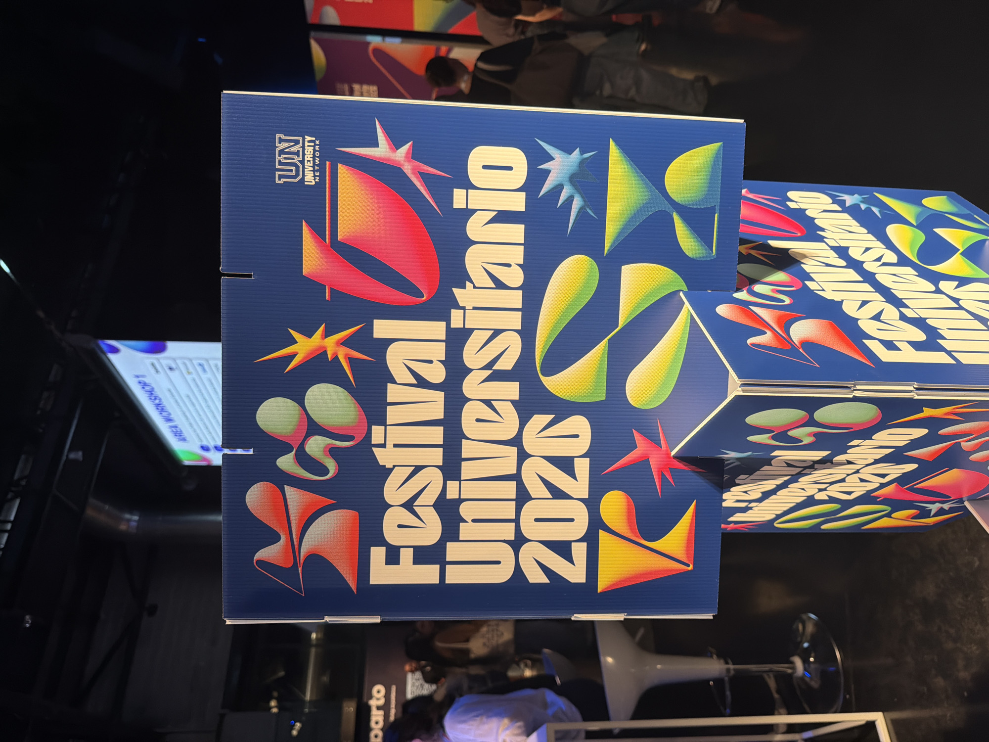 The image shows a close-up shot of blue cardboard signs with the words "Festival Universitario 2020" in white lettering, decorated with colorful abstract shapes and the logo of "UN University Network." The signs are angled and stacked on top of each other. The location is the city of Milan, Italy. The environment appears to be a dark, indoor space, possibly an exhibition or event area, as there are indistinct figures and objects in the background and reflections on the surfaces. The main subjects are the signs promoting the "Festival Universitario 2020."