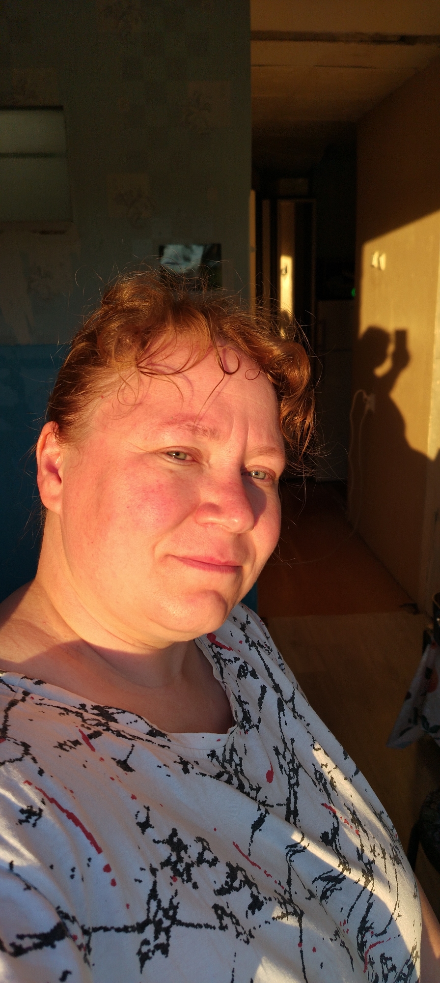The image shows a woman with reddish hair and a white shirt with black and red abstract patterns. She is facing the camera with a slight smile. The background includes a blue wall, a doorway, and a hallway with a light-colored wall. A shadow on the light-colored wall suggests someone holding a phone. The location is the city of Chaikovsky, Russia.