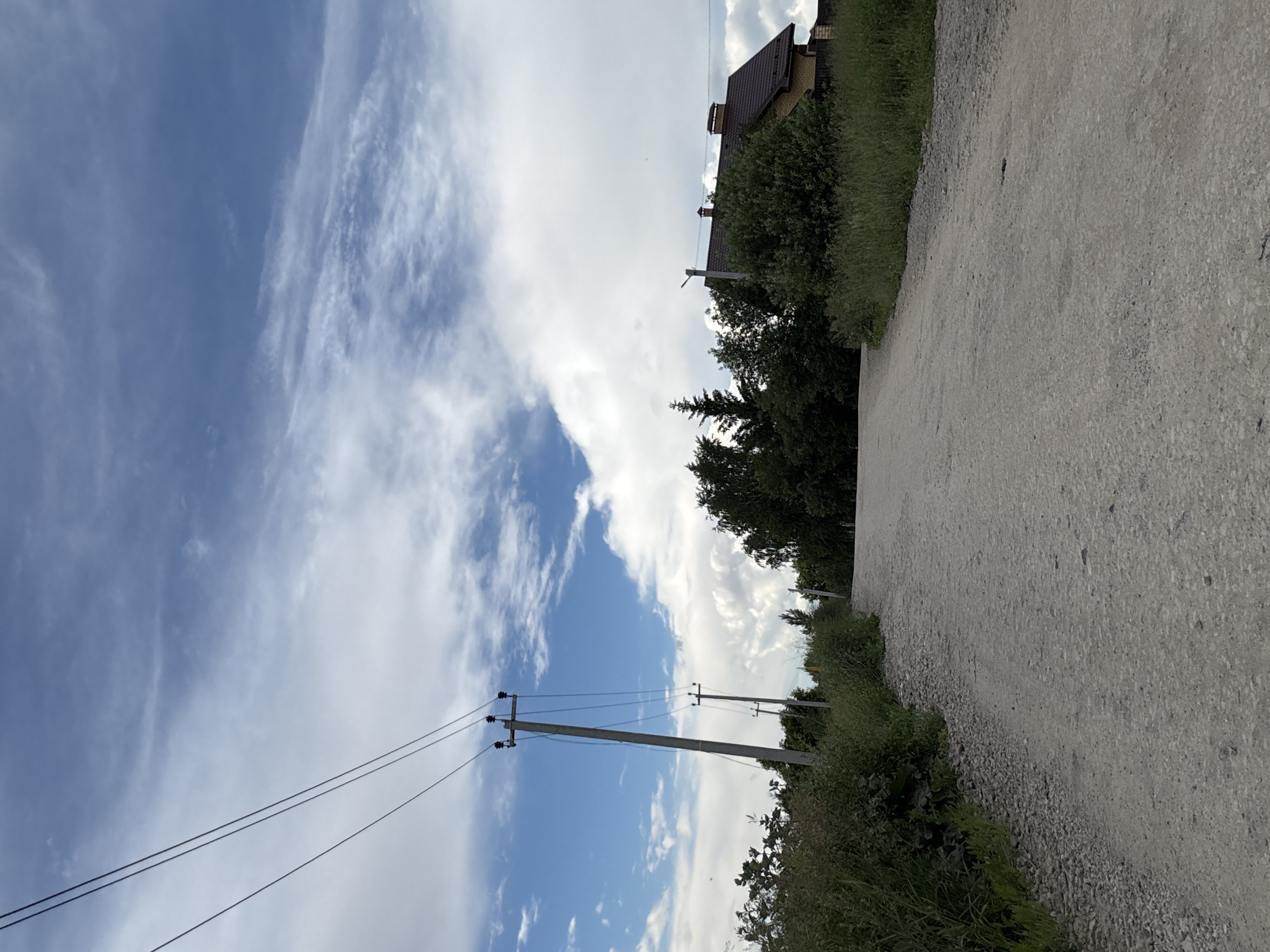 The image depicts a rural scene in Gorny, Russia, under a vast, blue sky with scattered white clouds. A dirt road stretches into the distance, bordered by lush green vegetation and trees. In the background, a house with a brown roof and a brick facade is visible, partially obscured by dense foliage. Electrical poles with wires run parallel to the road, hinting at the presence of infrastructure in this somewhat isolated setting. The overall atmosphere is peaceful and serene, suggesting a clear day with pleasant weather, likely late morning or early afternoon. There is no visible text in the image.