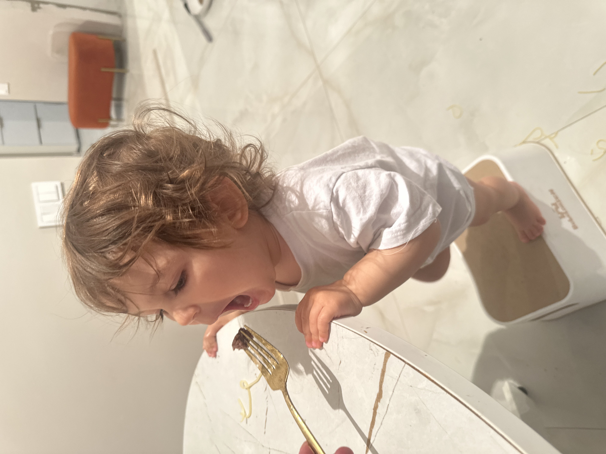 An eye-level indoor shot captures a young child, likely a toddler, enthusiastically engaging with food during mealtime in a home setting in Moscow, Russia. The child, with light brown, curly hair, wears a white short-sleeved t-shirt and leans forward, arms bent with hands placed on a light-colored, possibly marble-patterned table or high chair tray. The child's mouth is wide open, gazing intently at a gold-colored fork held by an unseen adult hand. A piece of dark-colored food, possibly meat, is on the fork, positioned just inches from the child's mouth.
Several strands of spaghetti are scattered across the table surface, suggesting a typical messy toddler meal. The floor beneath the child is tiled with a light, patterned material. Next to the child's bare feet is a white and beige child's learning tower or step stool. In the background, an orange modern stool or chair with gold legs is visible against a white wall, which also features light switches or outlets. Bright natural light illuminates the scene, casting distinct shadows from the fork and the child's head, indicating it is daytime. The child's expression conveys intense eagerness, anticipation, or hunger for the food being offered. No discernible text is visible in the image.