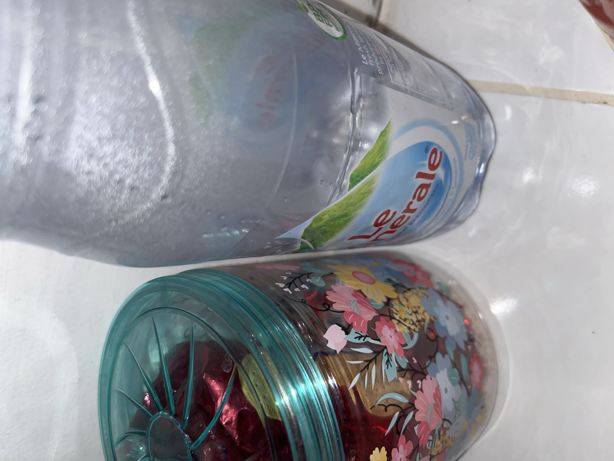 The image displays a still-life arrangement of two consumer items on a light-colored surface, situated in Lhokseumawe, Indonesia. On the right, a clear plastic bottle of "Le Minerale" mineral water is depicted, approximately two-thirds full, with condensation visible on its exterior. The label features a green landscape graphic and the text "Le Minerale," "pasumungan," and "BPA FREE." To the left, a transparent cylindrical jar with a teal, radially spoked plastic lid contains various food items; prominent among them are several pieces wrapped in reflective red foil, along with lighter, solid items. The lower section of the jar's exterior is decorated with a multi-colored floral motif, featuring pink, yellow, blue, and white blossoms alongside green foliage. Both containers rest on a white or light-grey tiled surface, exhibiting faint grout lines, against a uniform, light-colored background. No individuals are present.