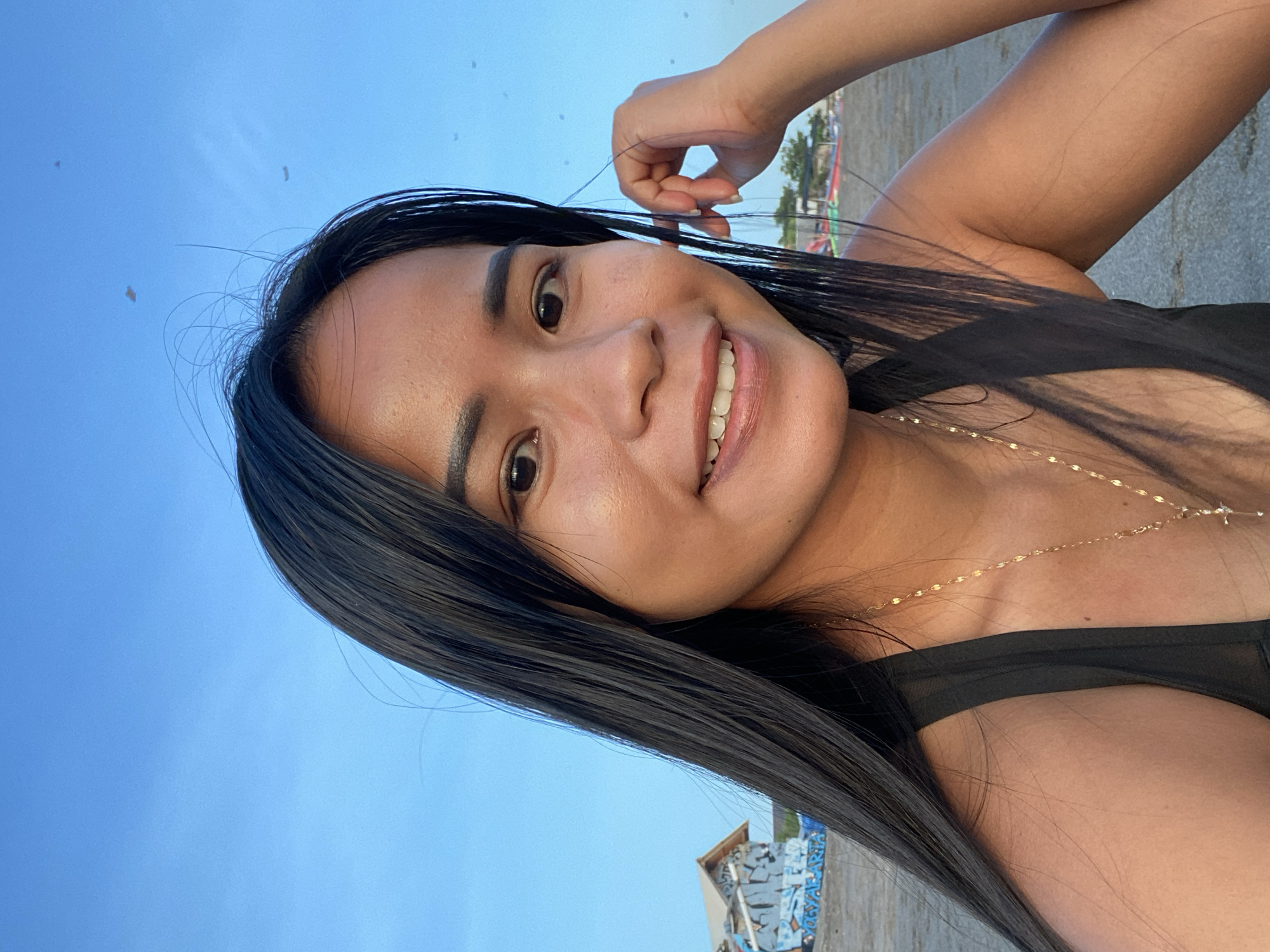 A woman with long, dark hair smiles directly at the camera. She is wearing a black top and a gold necklace. The background features a clear blue sky, a stretch of sandy beach, and a building with graffiti on it. The location appears to be in North Kuta, Indonesia.