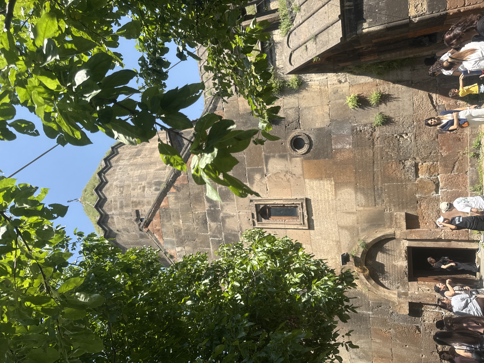 In Bjni, Armenia, a group of people stand before the ancient stone facade of a church, partially obscured by lush green tree branches against a clear blue sky. The church, constructed from rough-hewn reddish-brown stones, features a prominent arched entryway with a dark opening. Above the entrance, the weathered stone walls are adorned with inscriptions, and a small, rectangular window is visible to the left. A cylindrical opening is set into the wall to the right of the window, and a pointed structure with a cross on top, covered in grass, rises above the main structure. Some individuals are entering or exiting the church, while others observe its exterior. A young girl in a yellow shirt stands near a man wearing a light-colored cap and a white t-shirt. The scene is bathed in bright sunlight, casting shadows on the stone.