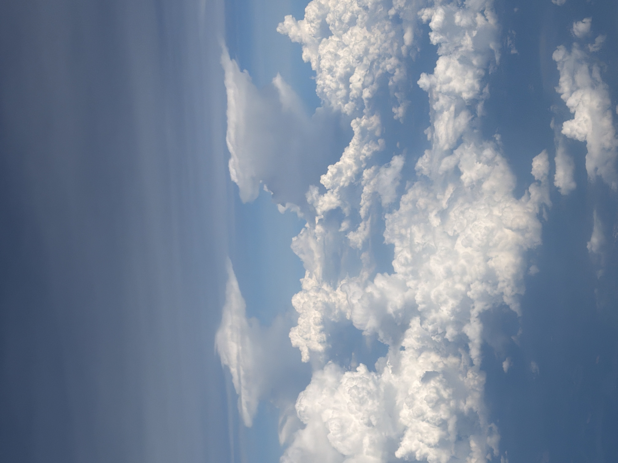 This image is an aerial view of clouds from an airplane. The clouds are white and fluffy, with some darker gray clouds visible in the distance. The sky is blue. There are no people, subjects, objects, actions, interactions, or city landmarks visible in this image. The image does not depict Hong Kong, China or any specific location.