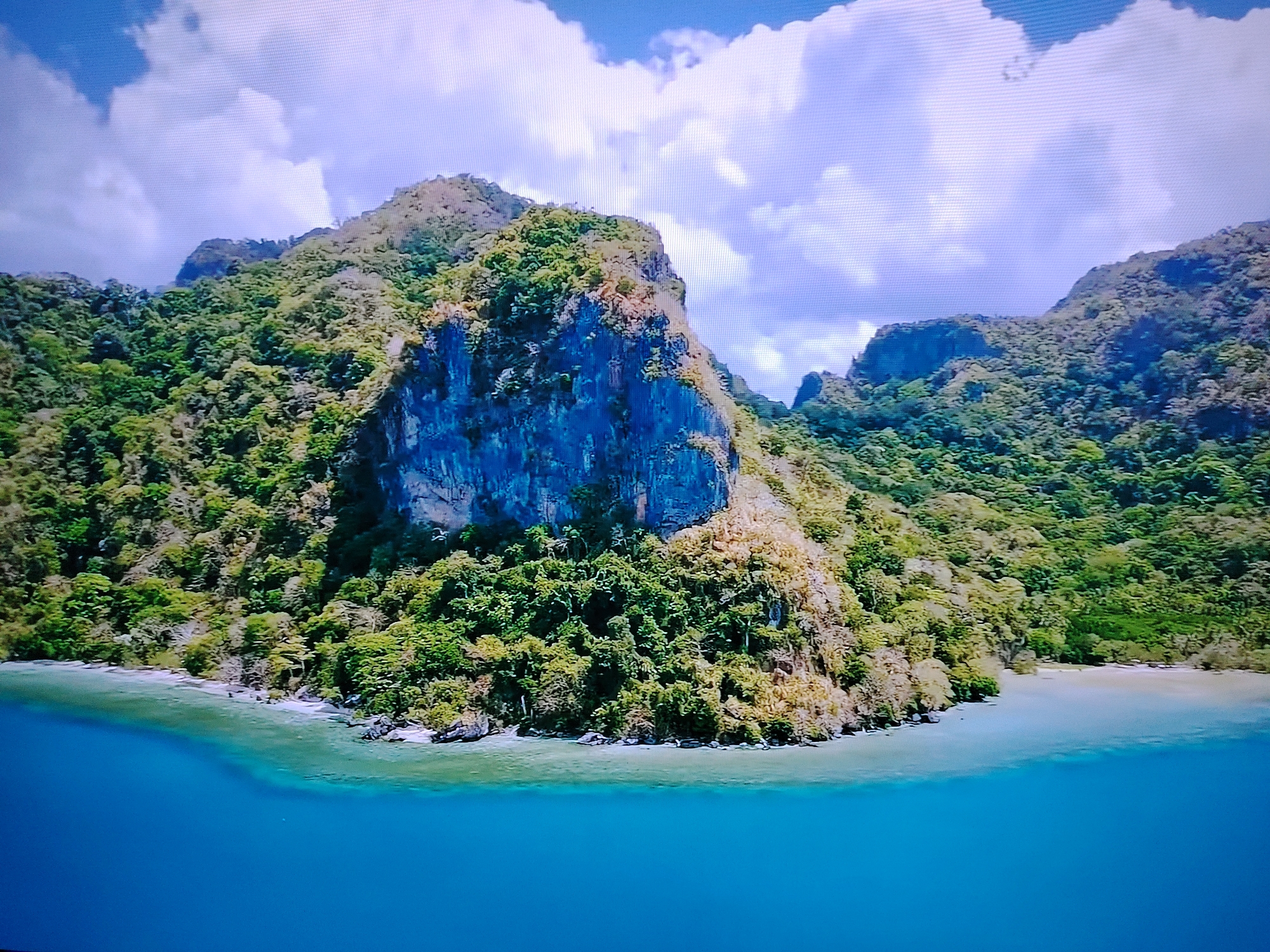 The image depicts a tropical island landscape. A large, rugged, mountainous island covered in dense green foliage dominates the scene. The island features a sheer, rocky cliff face with a distinct blue hue. The shoreline is a narrow strip of sand and rocks, meeting crystal-clear turquoise water. The water transitions into a deep blue expanse in the foreground. In the sky, large, fluffy white clouds drift against a pale blue background. The scene appears to be a natural, undeveloped coastal environment. There is no indication of the city of Huntingdonshire, United Kingdom, in this image.