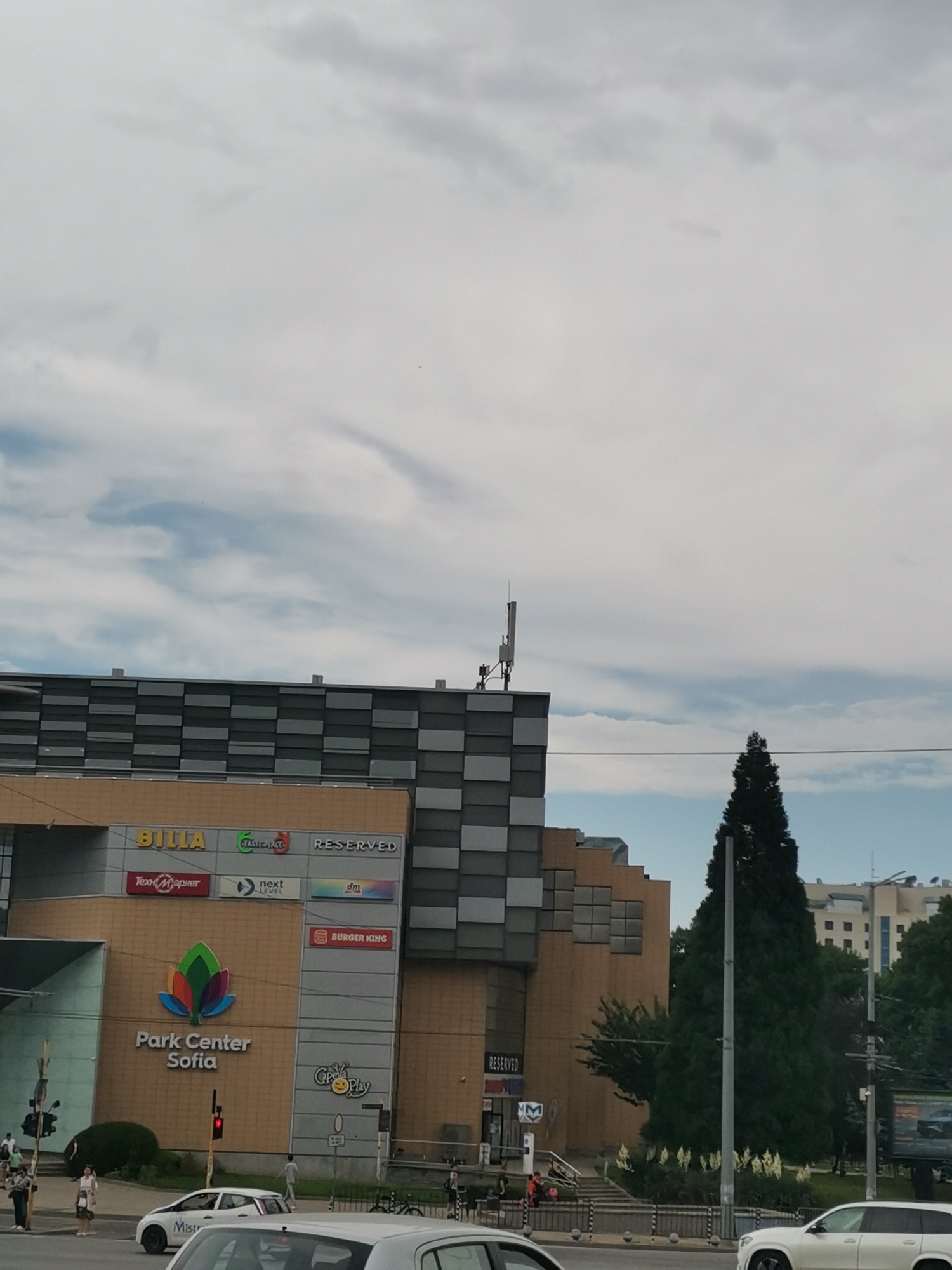 This image depicts the Park Center Sofia shopping mall, located in Sofia, Bulgaria. The mall features a modern architectural design with a facade composed of tan and gray geometric panels. Prominent signage identifies the mall with "Park Center Sofia" and its colorful floral logo. Various retail brands are visible on the building's exterior, including BILLA, Reserved, dm, and Burger King.
In the foreground, a street scene unfolds with several vehicles, including a white sedan and a white SUV. Pedestrians are scattered along the sidewalk and crossing the street. Traffic lights are present, with one showing a red signal. The overall environment suggests a bustling urban setting on an overcast day. A tall, dark green evergreen tree stands prominently to the right of the mall, adding a natural element to the cityscape. Buildings and trees are visible in the background, extending into the distance.