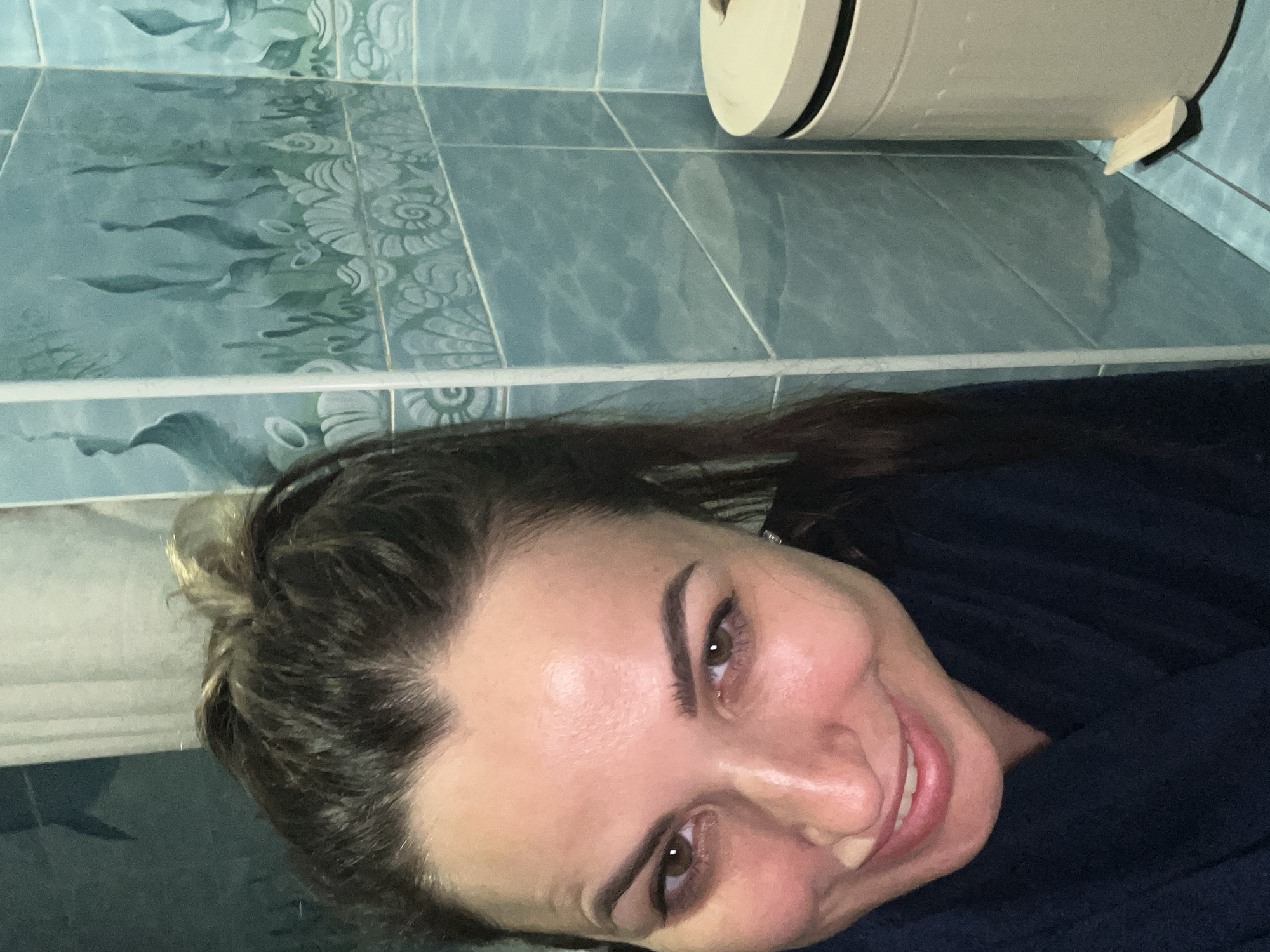 The image is a close-up selfie of a woman in a bathroom. She is looking directly at the camera with a smile. Her hair is pulled back, and she is wearing a dark blue robe. The bathroom walls are tiled with a blue, underwater-themed pattern, featuring shells and seaweed. A light-colored trash can with a foot pedal is visible on the right side of the frame. The lighting suggests the image was taken indoors, likely in a residential bathroom.