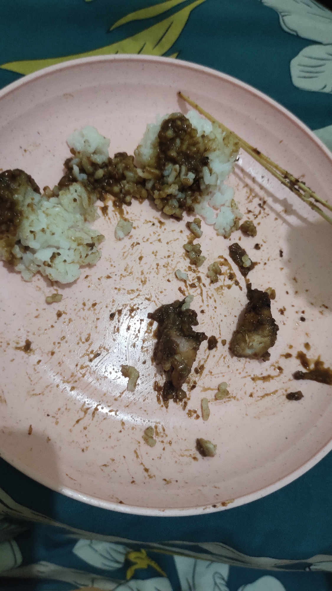 A pink plate with leftover food and a wooden skewer is on a table covered with a patterned cloth. The plate contains white rice mixed with a dark sauce, and some pieces of meat. The food appears to have been eaten. There are no people visible in the image. The background shows a teal cloth with a yellow floral pattern. The location is the city of Kuwarisan, Indonesia.