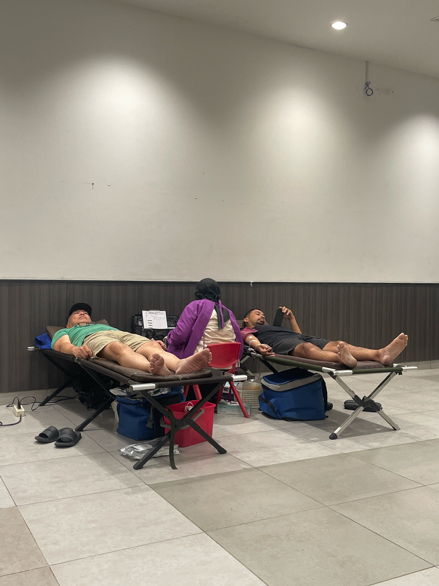 Two men are lying on cots, appearing to be donating blood in a facility in Pagedangan, Indonesia. A person wearing a purple top and a headscarf is attending to one of the men. The room has light-colored walls with ceiling lights, and the floor is tiled. Various items like bags, a red bucket, and medical equipment are visible around the cots. The men are dressed casually in t-shirts and shorts. The overall environment suggests a medical or donation center.