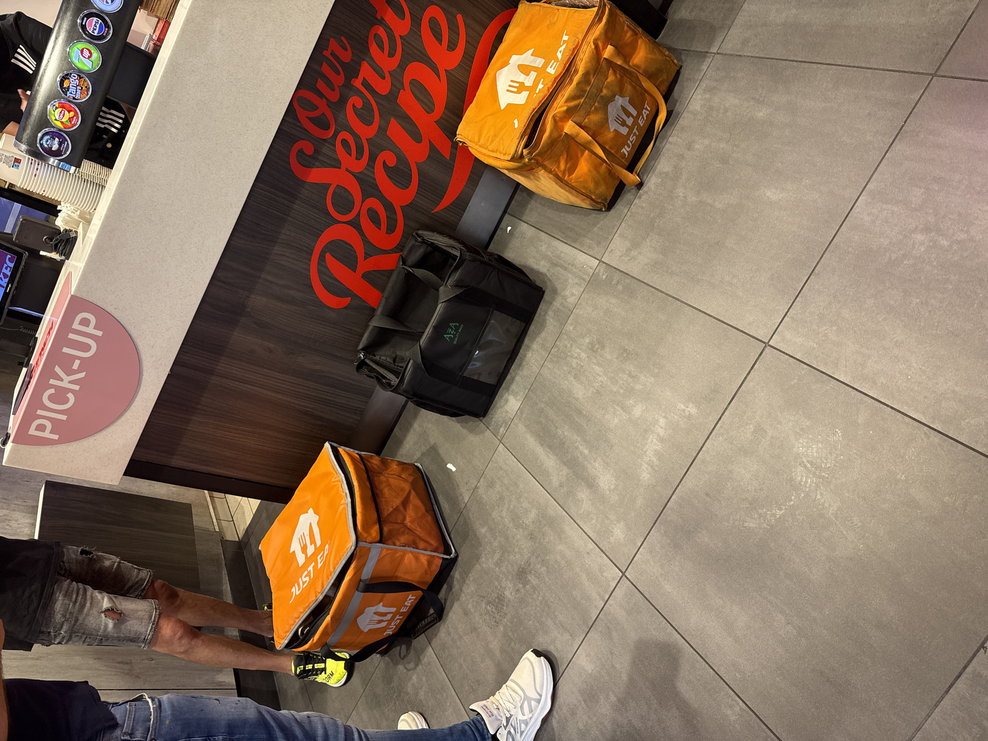 The scene is inside a KFC restaurant in Great Yarmouth, United Kingdom. Two delivery drivers are present with their delivery bags. The bags are resting on the floor. One is wearing a black shirt, ripped jean shorts, and neon yellow and black sneakers. The second delivery driver is wearing blue jeans and white sneakers. There are three delivery bags in total. Two orange bags bearing the "JUST EAT" logo are visible, one in the upper right corner of the frame, and one at the bottom left of the frame, next to the second delivery driver. There is also a black delivery bag with the "AZA QUALITY BAGS" logo, resting in the center of the frame. Behind the bags is a KFC counter, decorated with the slogan "Our Secret Recipe". To the left of the counter is a drink dispenser, a "PICK-UP" sign, and a computer screen with the letters "KEC" visible on the display. The floor is covered in gray square tiles.
