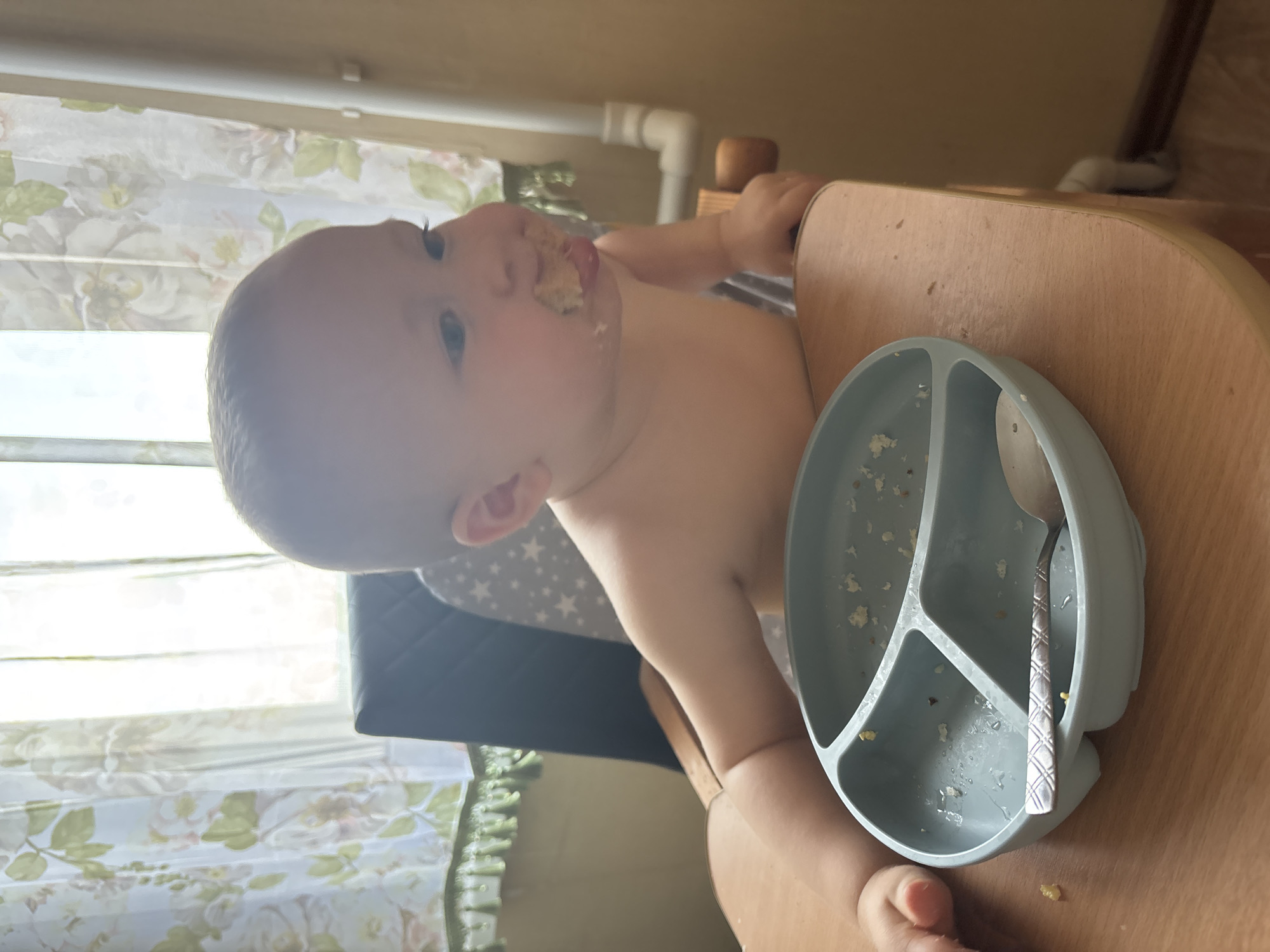 A baby is sitting in a high chair, facing left. The baby has food on its face and tongue, suggesting it is in the process of eating. In front of the baby is a light blue suction plate with three compartments, one of which contains a spoon. The plate and spoon appear to have some food residue on them. The high chair is made of wood, and the baby is wearing a grey bib with white stars. In the background, a window with floral curtains is visible. The overall scene suggests a mealtime for a young child. The image was taken in Naberezhnye Chelny, Russia. The time of day is not explicitly stated, but the lighting suggests it is daytime.