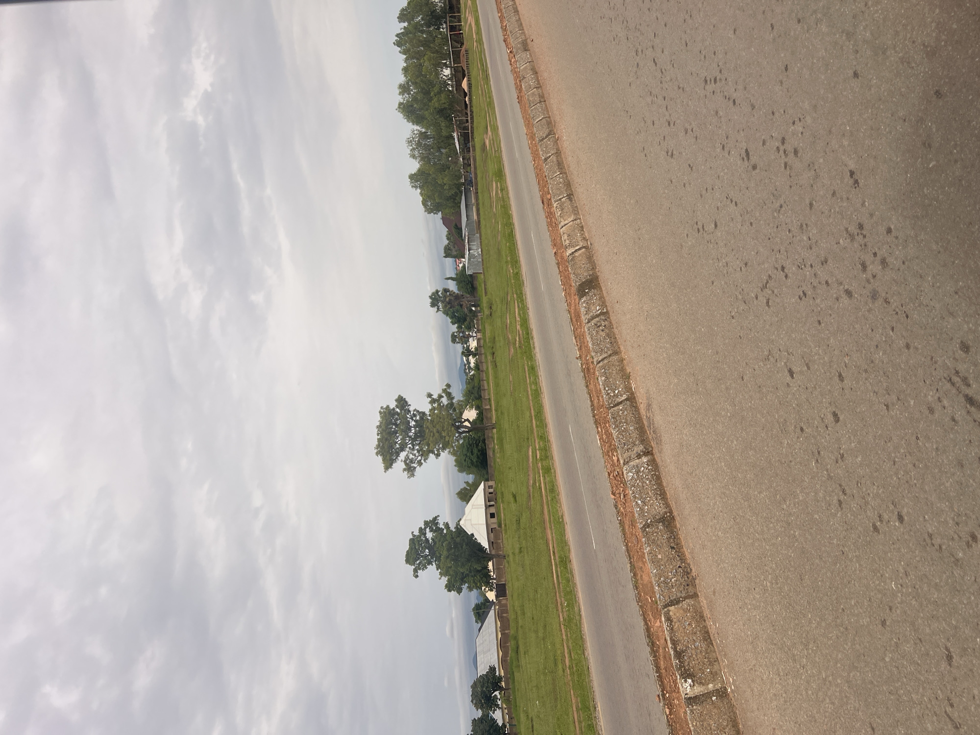The image depicts a roadside scene in Jalingo, Nigeria. The perspective is from a slightly elevated, angled viewpoint looking down a paved road that curves to the right. The road appears to be asphalt, with some dark markings or stains visible. It is bordered by a raised curb made of concrete blocks and what appears to be dirt.
To the left of the road is a strip of vibrant green grass, suggesting a well-maintained area alongside the thoroughfare. Beyond the grassy verge, several buildings are visible, some appearing to be under construction with exposed brickwork and unfinished roofs, while others seem complete with tiled roofs. Among the buildings, numerous trees with lush green foliage are scattered, some large and mature, others smaller. A few palm trees are also present.
The sky is overcast with soft, grey clouds, indicating a possibly humid or rainy day, or perhaps just a cloudy afternoon. The overall lighting suggests it is daytime, but the cloud cover diffuses the sunlight. There are no people visible in the frame, and the scene appears to be quiet and relatively undeveloped, with a mix of residential structures and natural vegetation. The combination of the road, grass, buildings, and trees points to a semi-urban or suburban setting. No text is visible in the image.